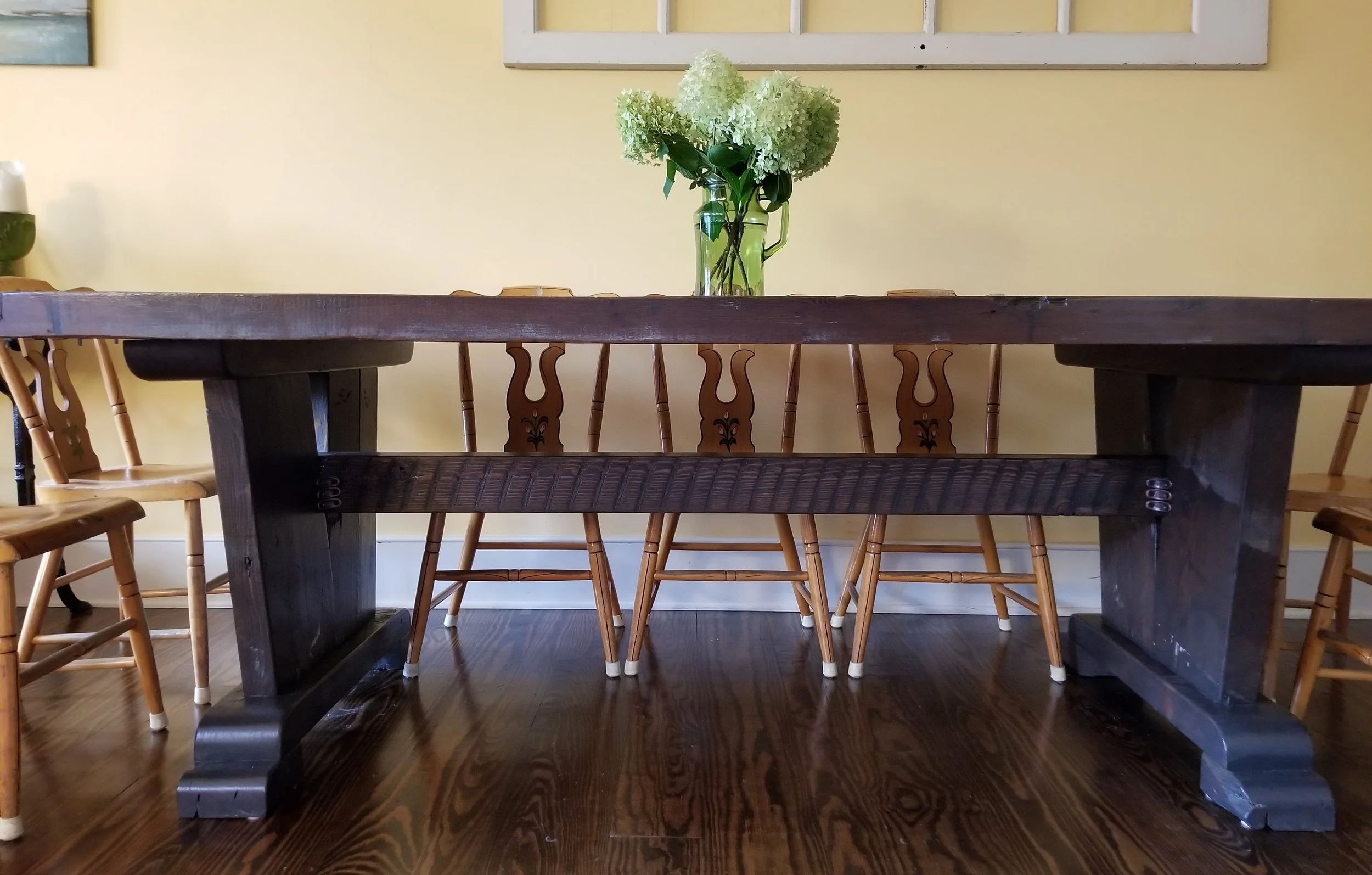 A wooden dining table with a dark finish, surrounded by wooden chairs with light-colored seats. A vase with white hydrangea flowers sits at the center of the table. The background features a yellow wall with a window frame and part of a painting visi