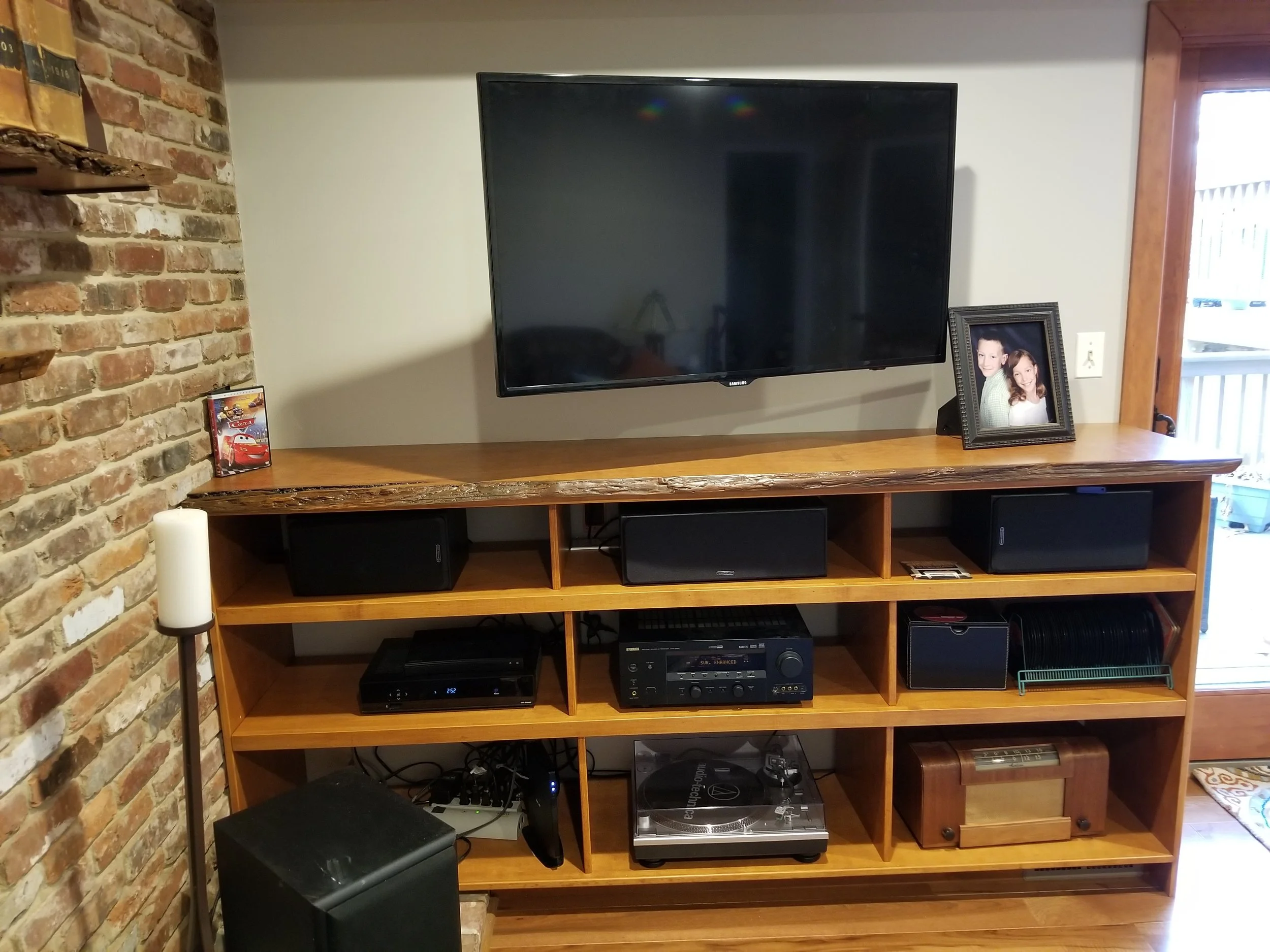 A custom built living room entertainment center with a large cherry live edge top. To the right of the TV, there is a framed photo of two children. To the left, a small framed picture of Lightning McQueen from Cars. In the entertainment cabinet are v