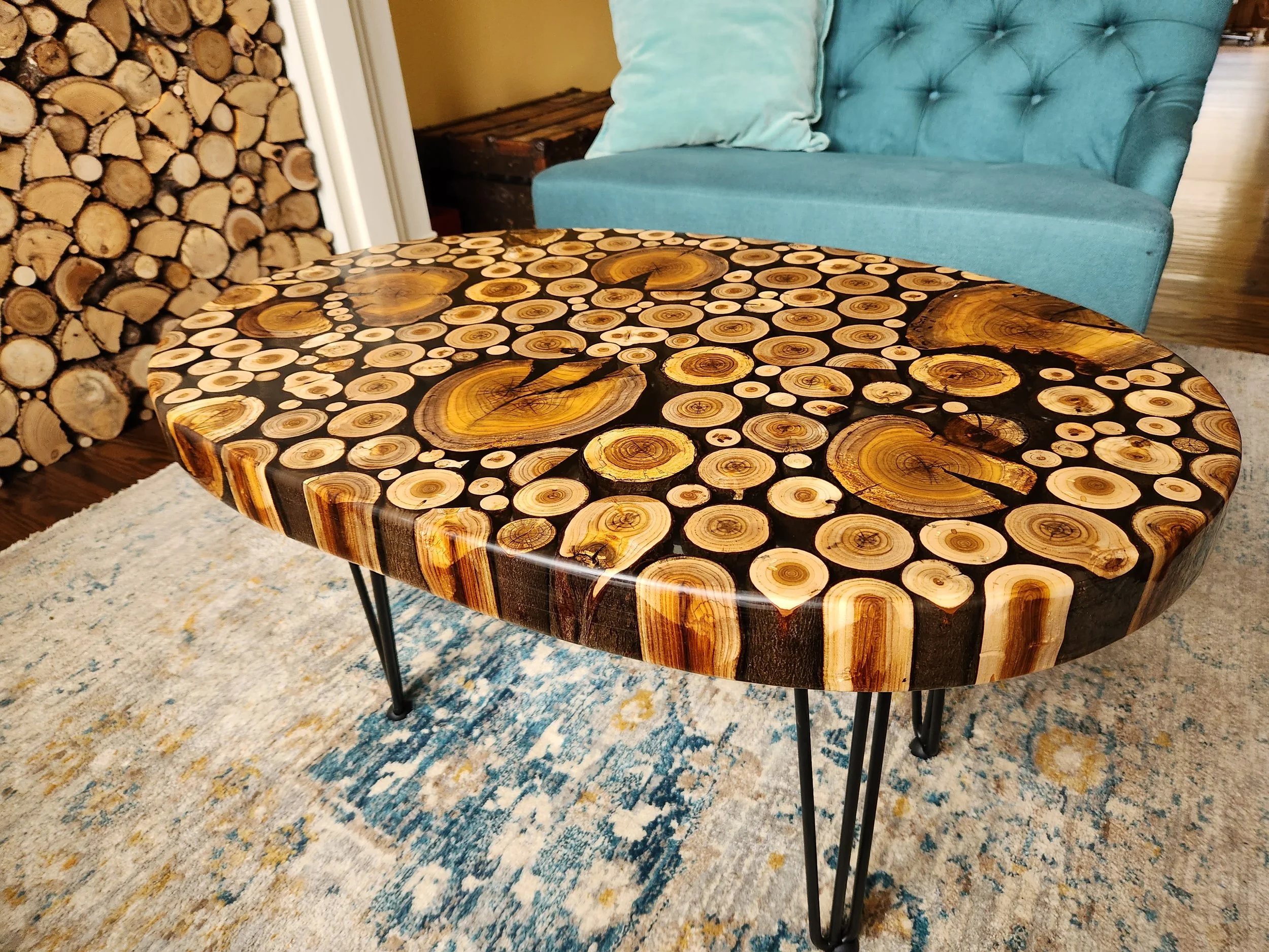 An indoor living room with a patterned custom poured clear epoxy table, a blue sofa with pastel pillows, a pile of chopped wood stacked against the wall, a rustic wooden storage chest, a colorful rug, and hardwood flooring.