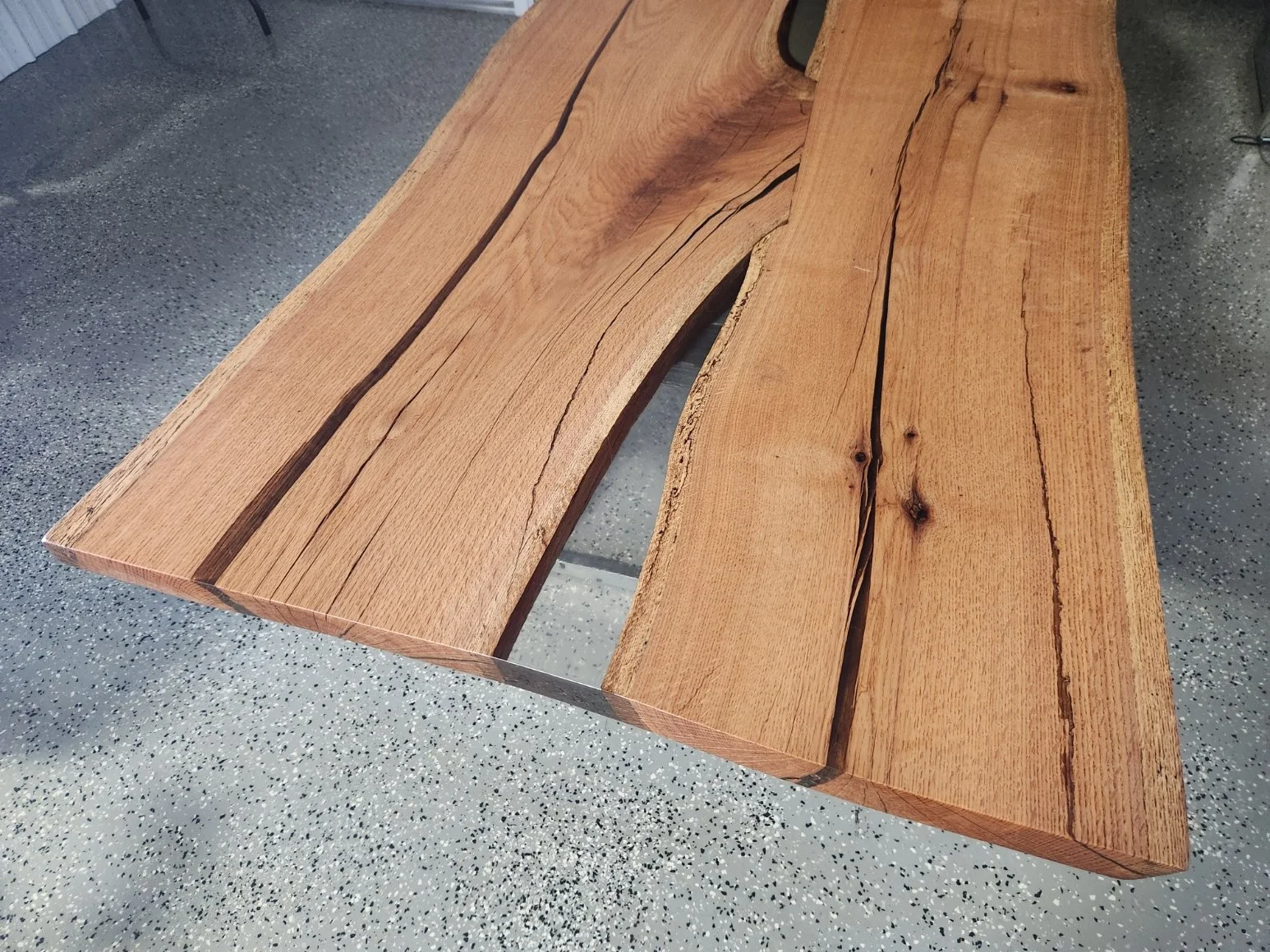 Close-up view of a wooden conference table made from thick, natural wood planks with visible grain patterns and cracks filled with clear epoxy, placed on a speckled gray concrete floor.