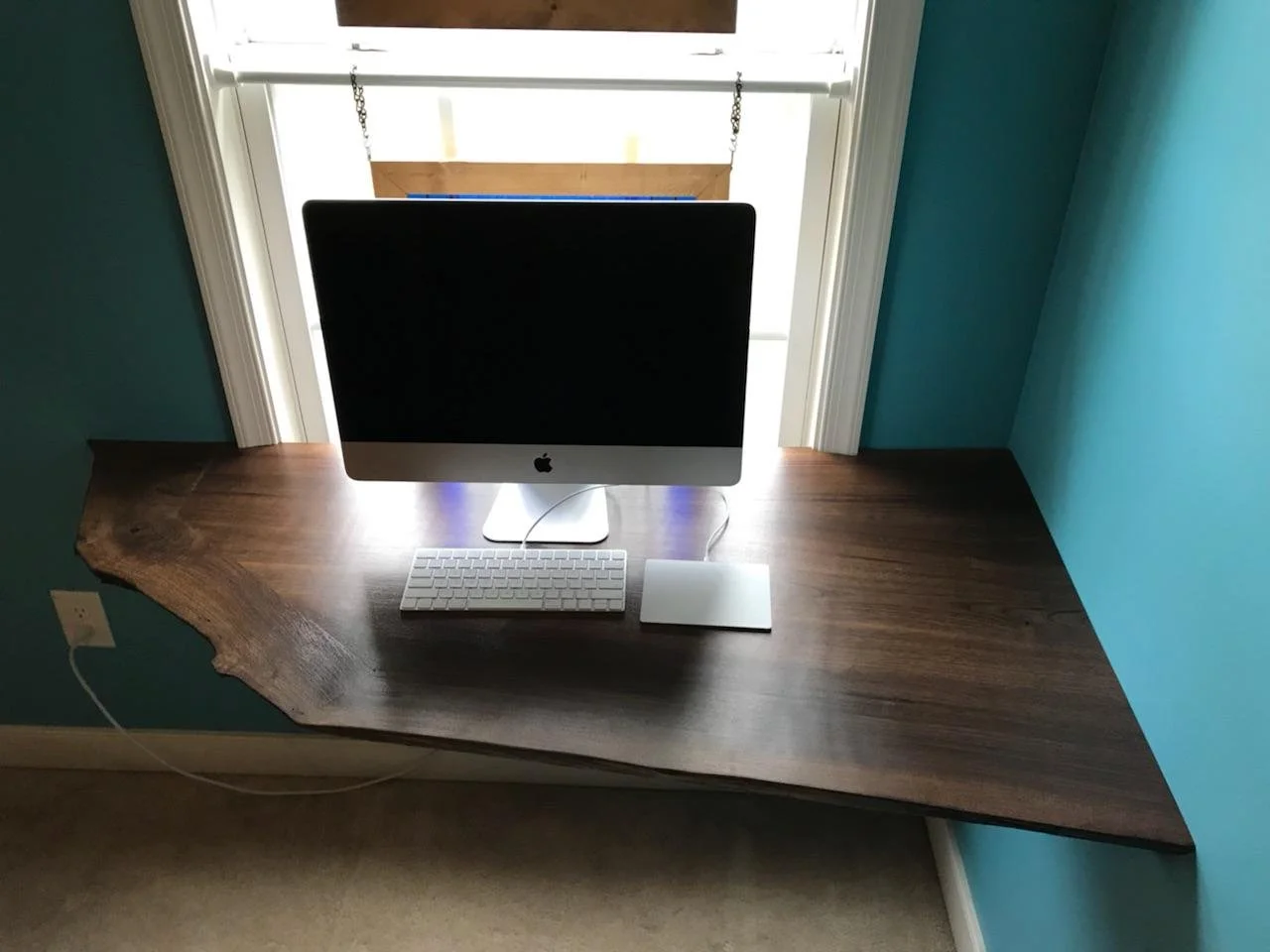 A walnut desk with a live edge finish holding an iMac computer, a white keyboard, and a mouse, situated in front of a window with wooden shutters, against teal walls.