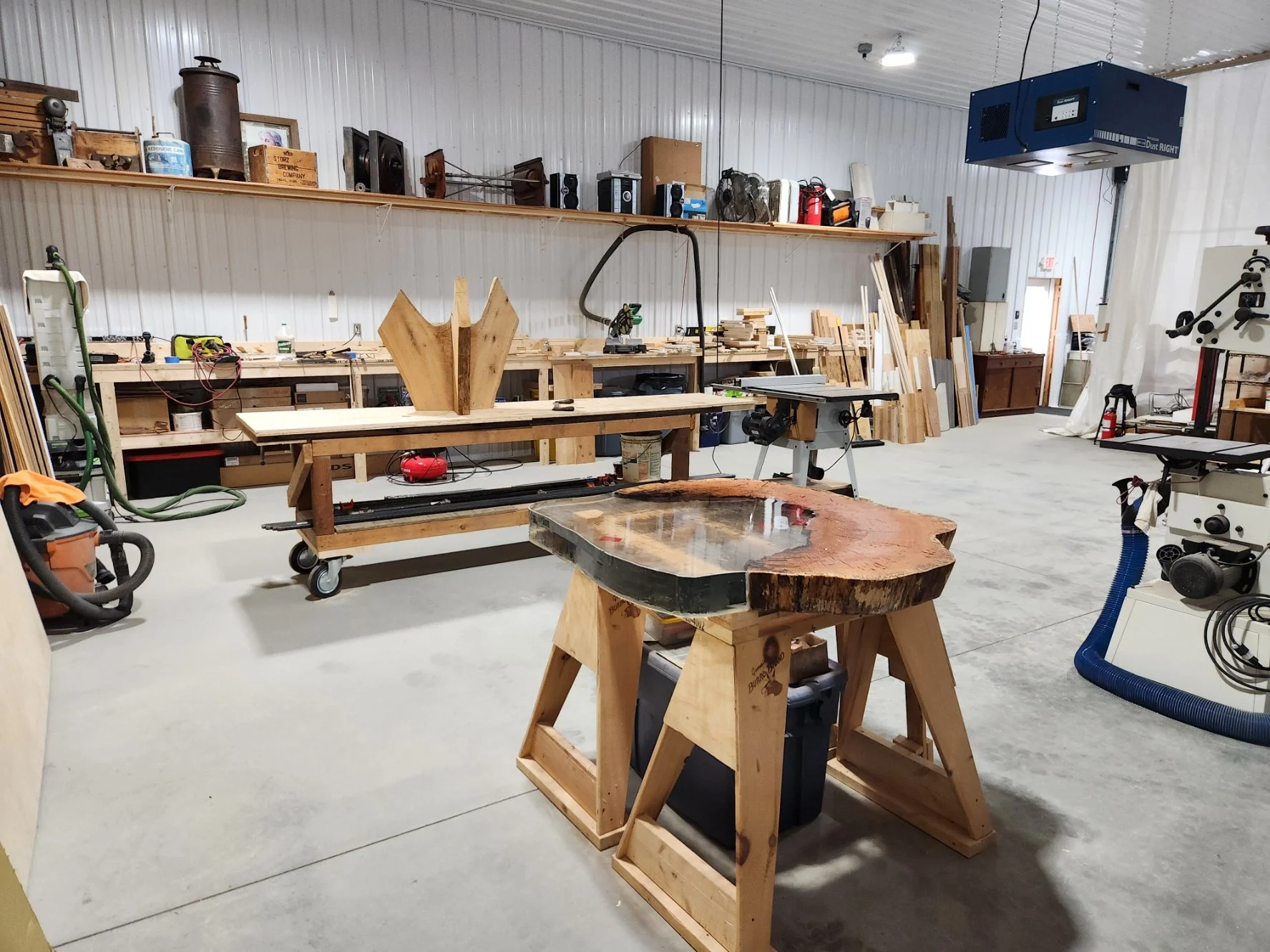 A woodworking workshop with various tools, wood pieces, and equipment including a table saw, workbenches, and storage shelves in a spacious garage-like area.
