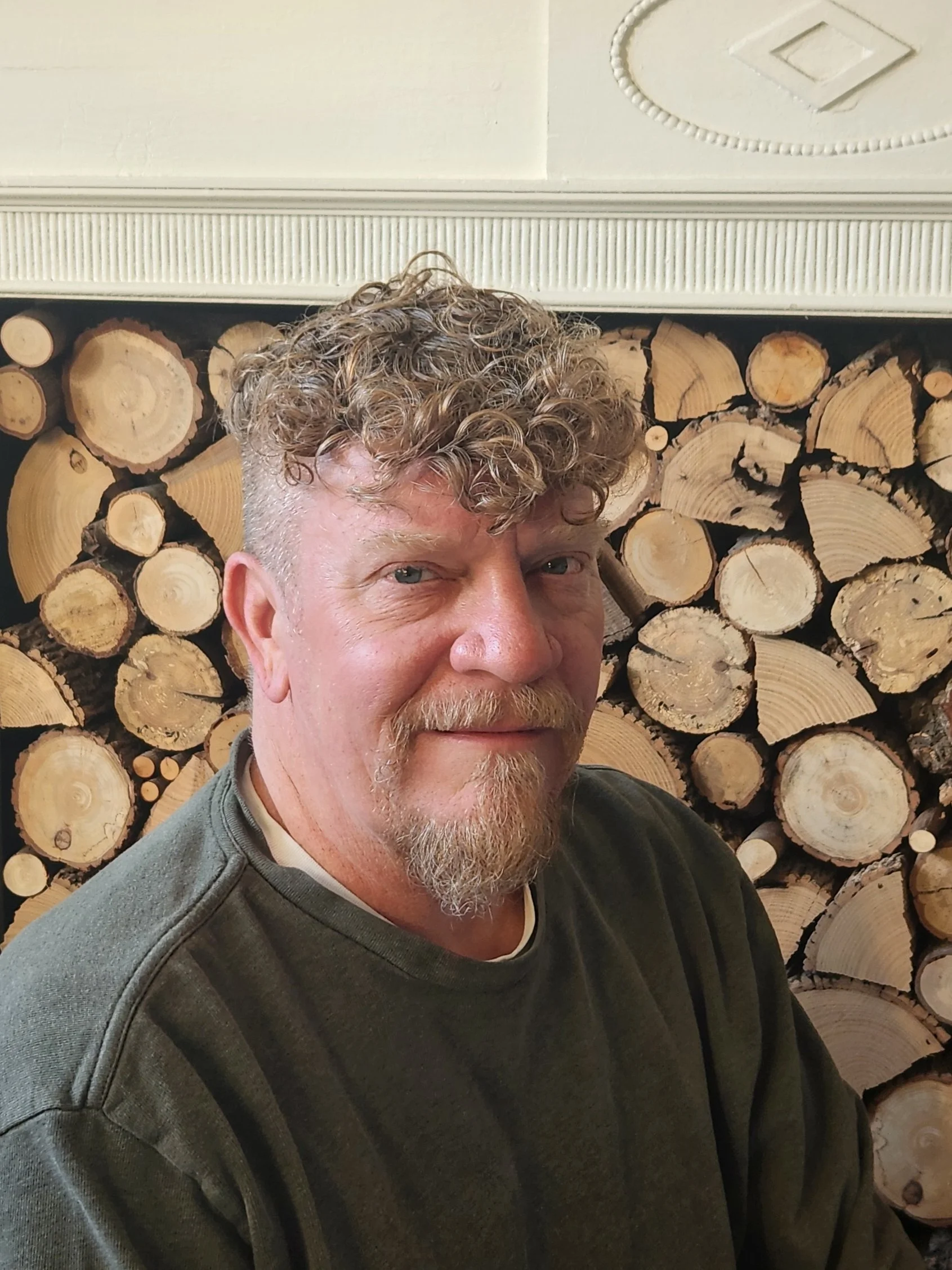 A man with curly light brown hair, a beard, and wearing a dark green hoodie sitting in front of a wall stacked with cut logs.
