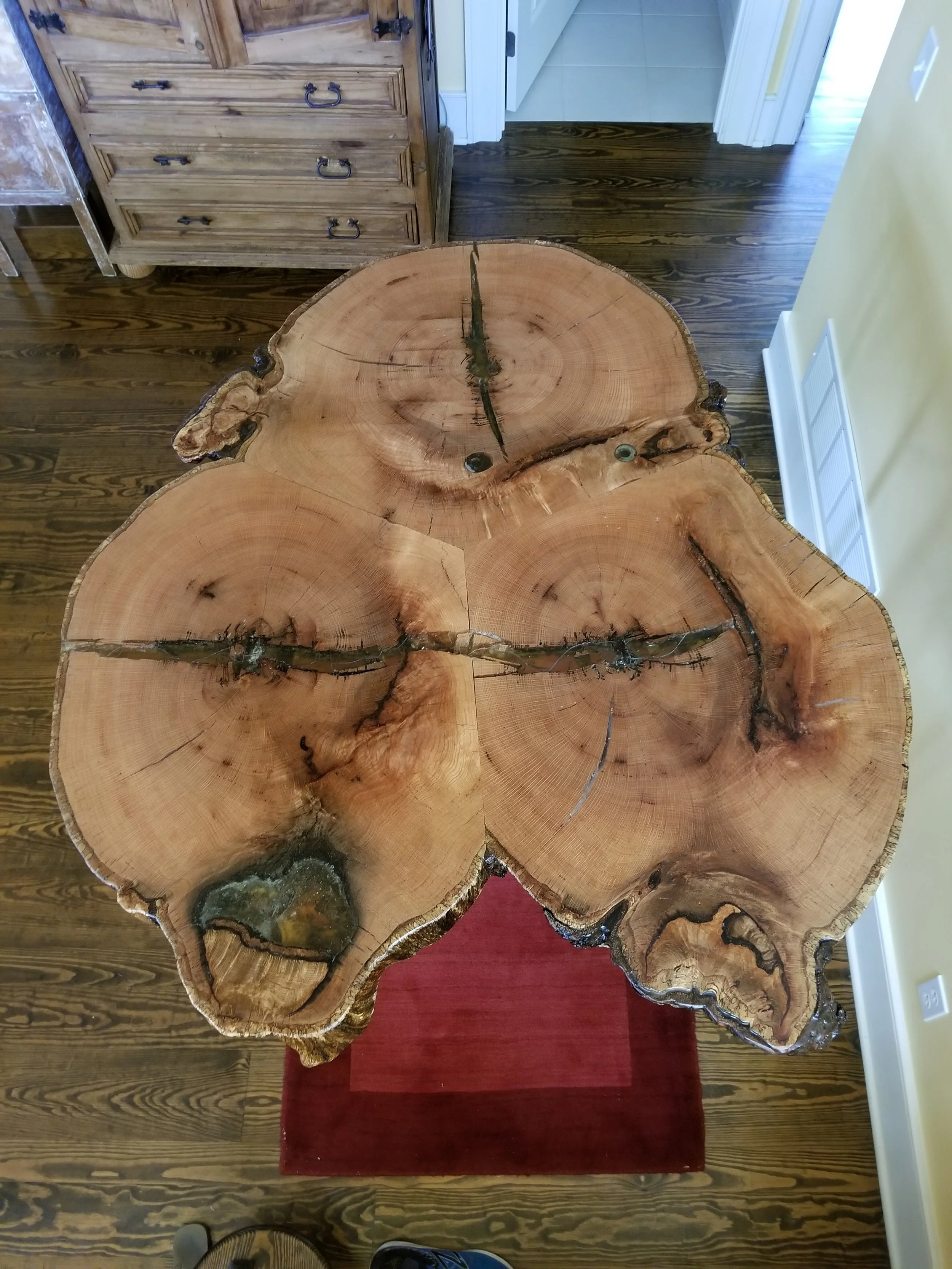 A natural wood high top table made from a large wood cross section with visible growth rings and cracks, placed on a red rug in a room with wooden flooring and a yellow wall.