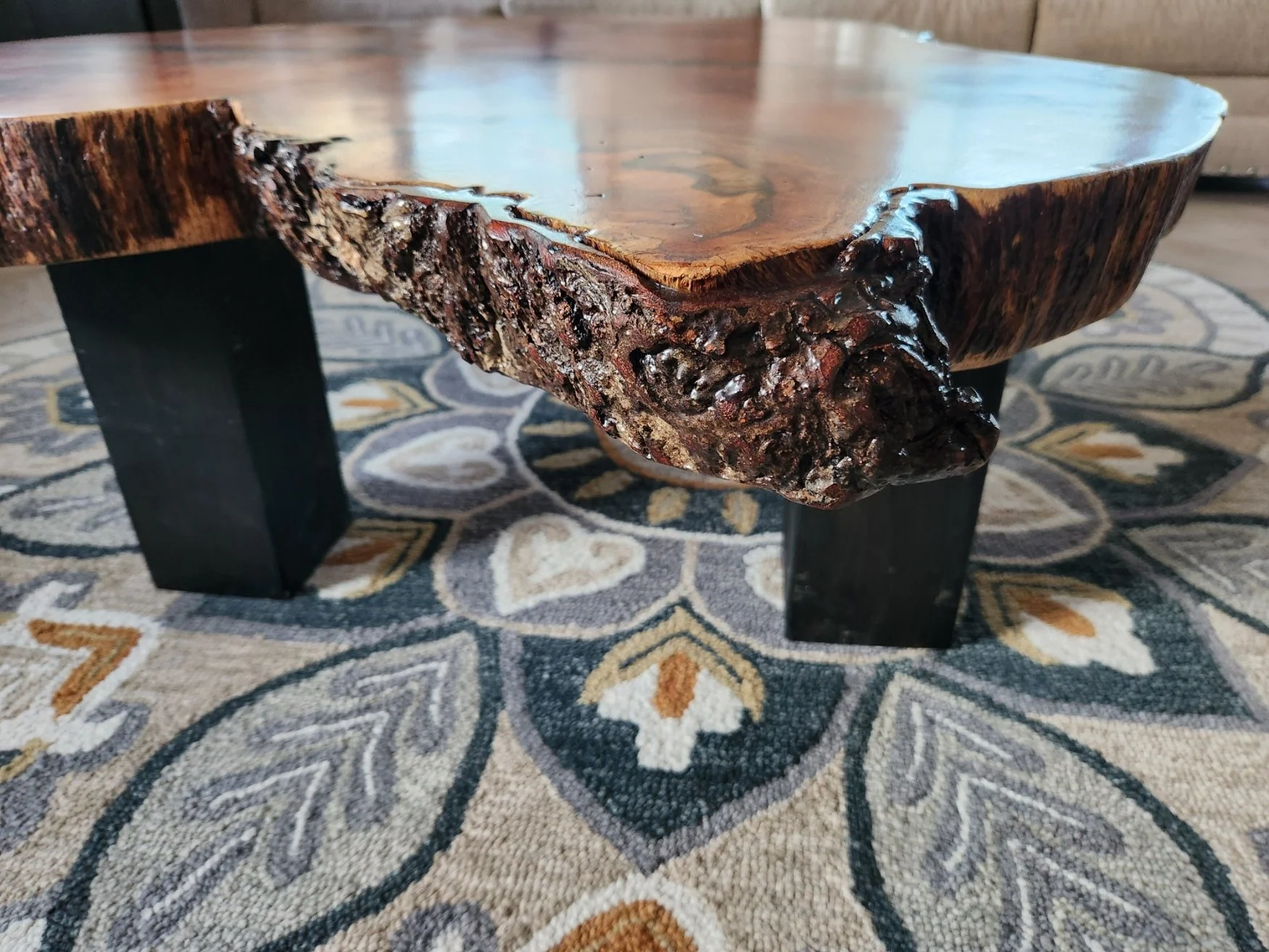 A wooden coffee table with a thick, polished natural wood top and dark, solid legs, placed on a patterned area rug.