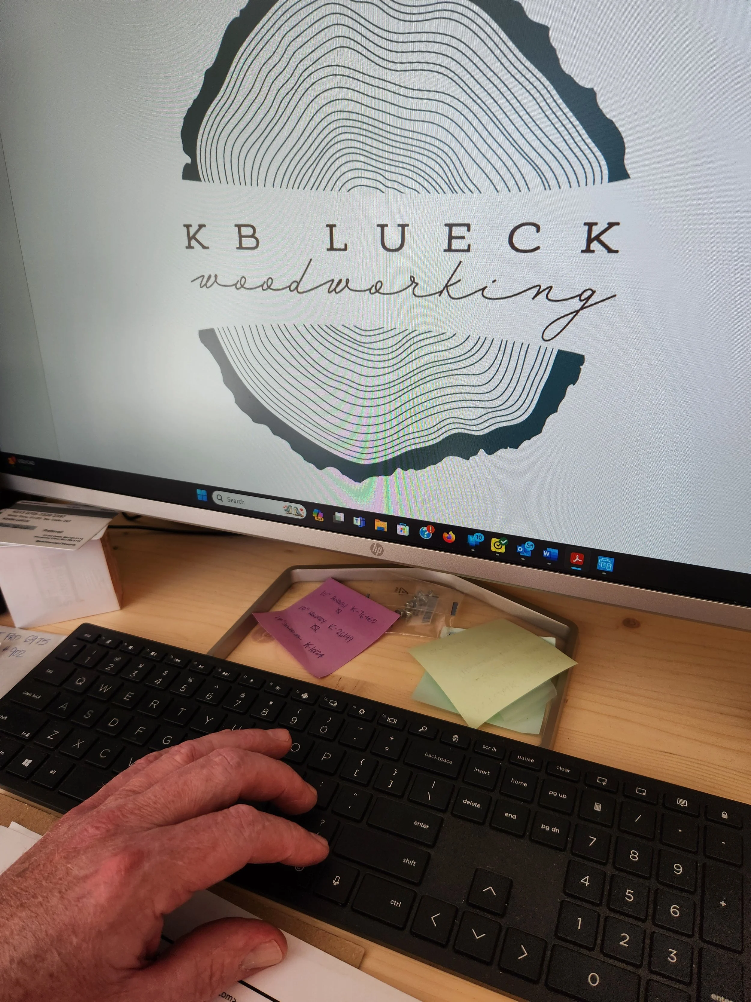 A computer monitor displaying a logo for KB Lueck Woodworking with a wooden circle with lines resembling tree rings. A person's hand is on a black keyboard in front of the monitor, and the workspace has sticky notes and office supplies.