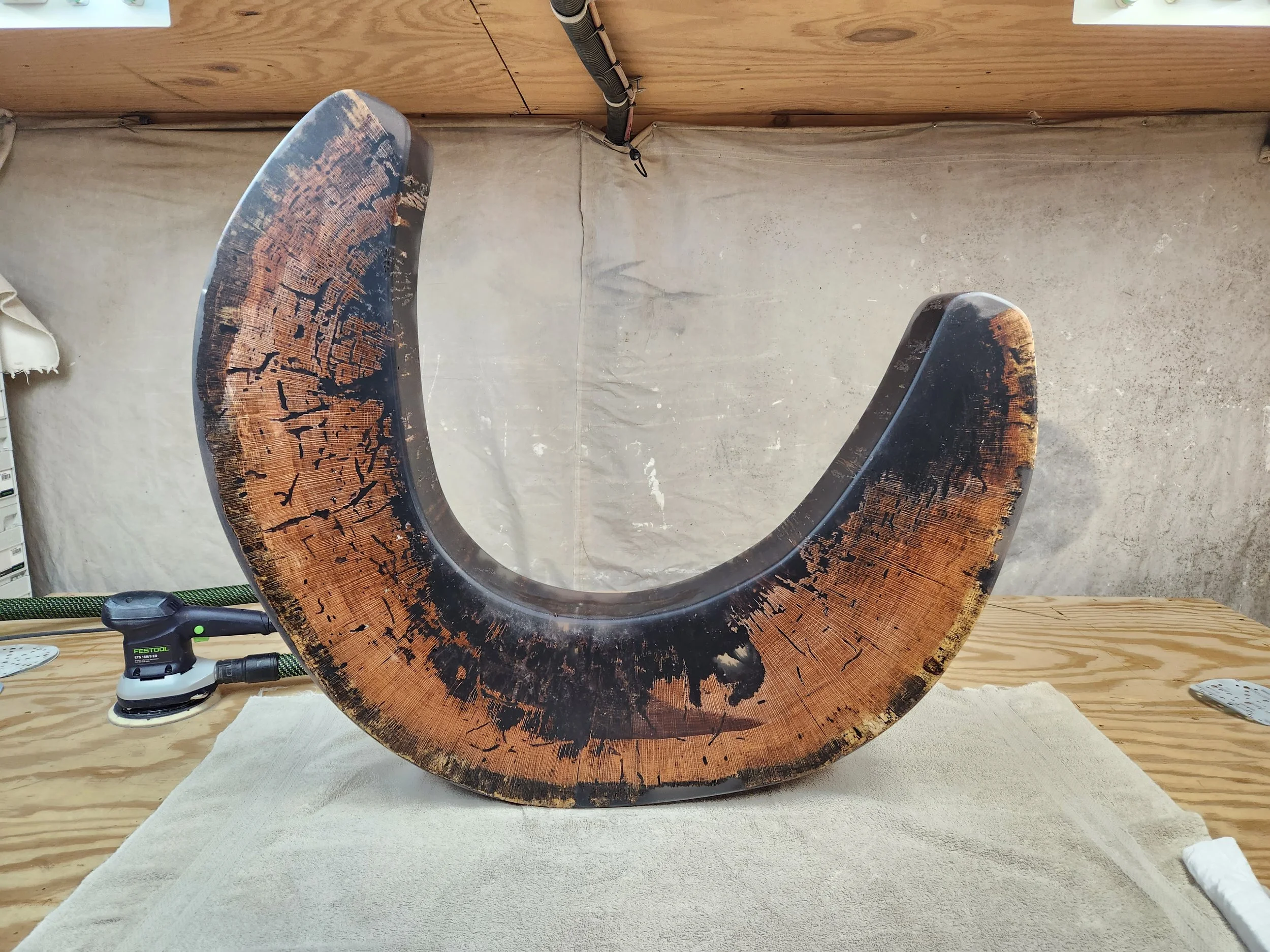 A large, curved wooden sculpture with a burnt pattern on its surface is displayed on a white cloth on a wooden table. Tools and a metal clamp are visible near the sculpture, with a concrete wall and wooden panel in the background.