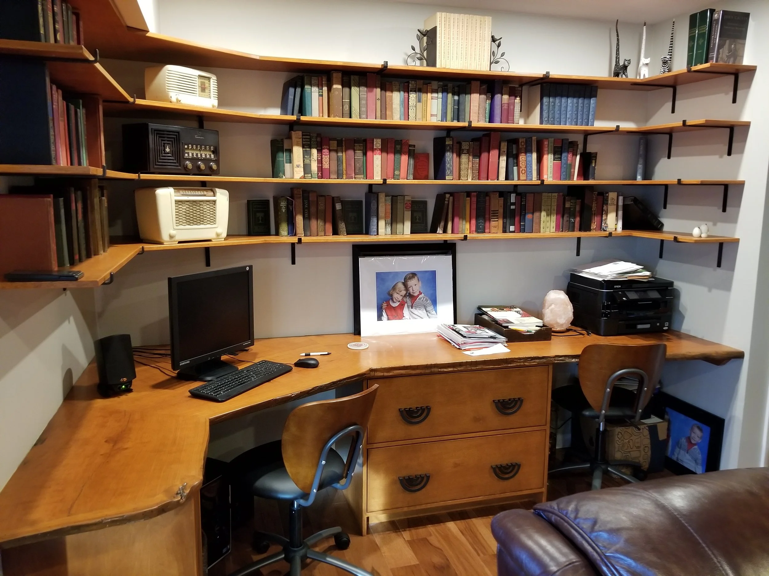 A home office with custom built cherry live edge desk with pull out file cabinet, black monitor, desk lamp, computer mouse, keyboard, and stacks of papers. Behind it, there are three custom built cherry bookshelves filled with books, and decorative i