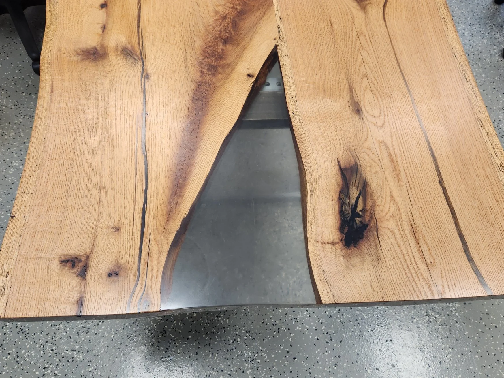 Close-up of a rustic wooden conference table with a live edge, showing natural wood grain, knots with clear epoxy on a speckled gray concrete floor.