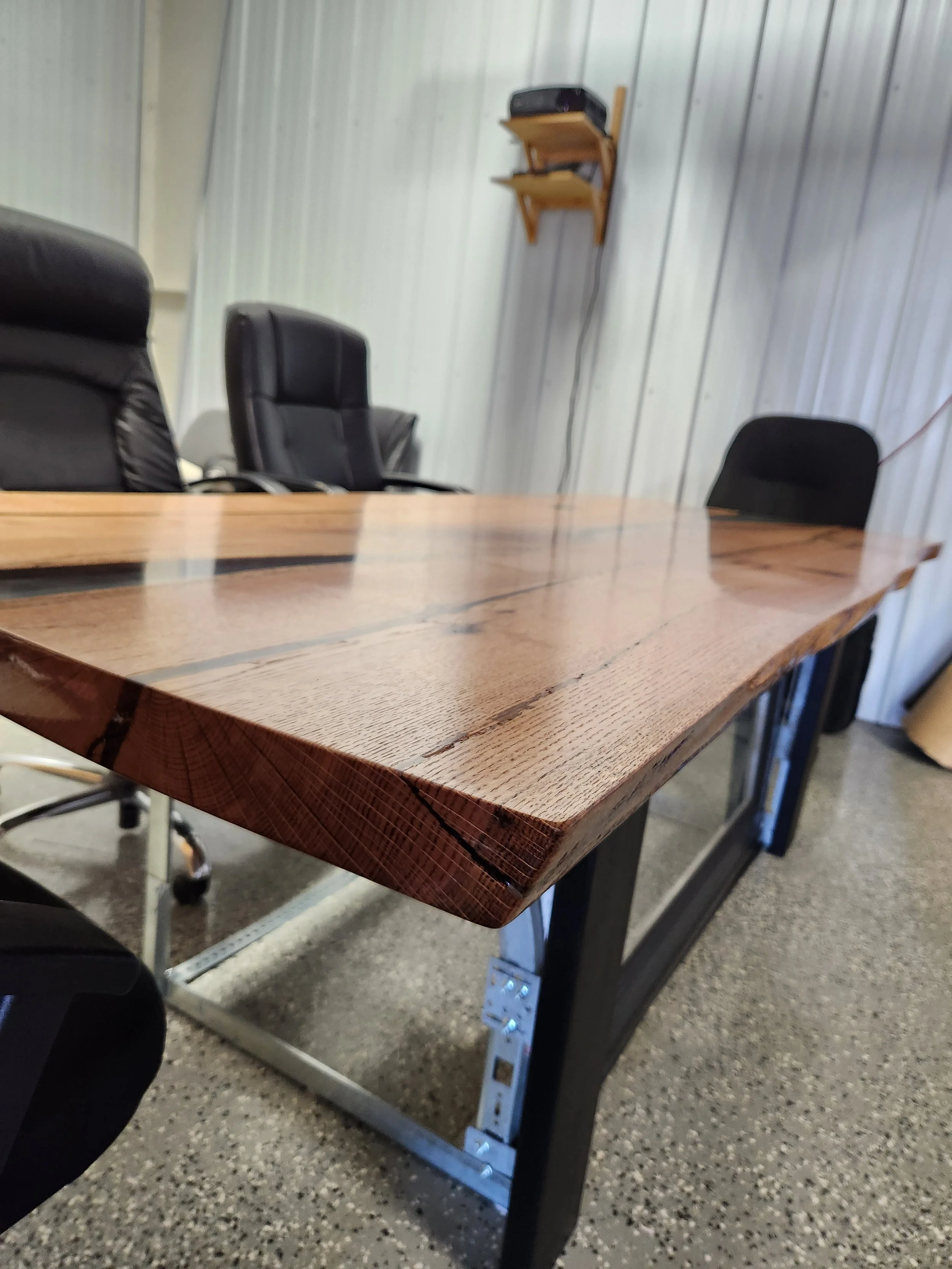 Live edge oak wooden table with metal legs and mini garage door panel in the center.  Surrounded by black office chairs in a room with metal paneled walls and a shelf with a projector on it.
