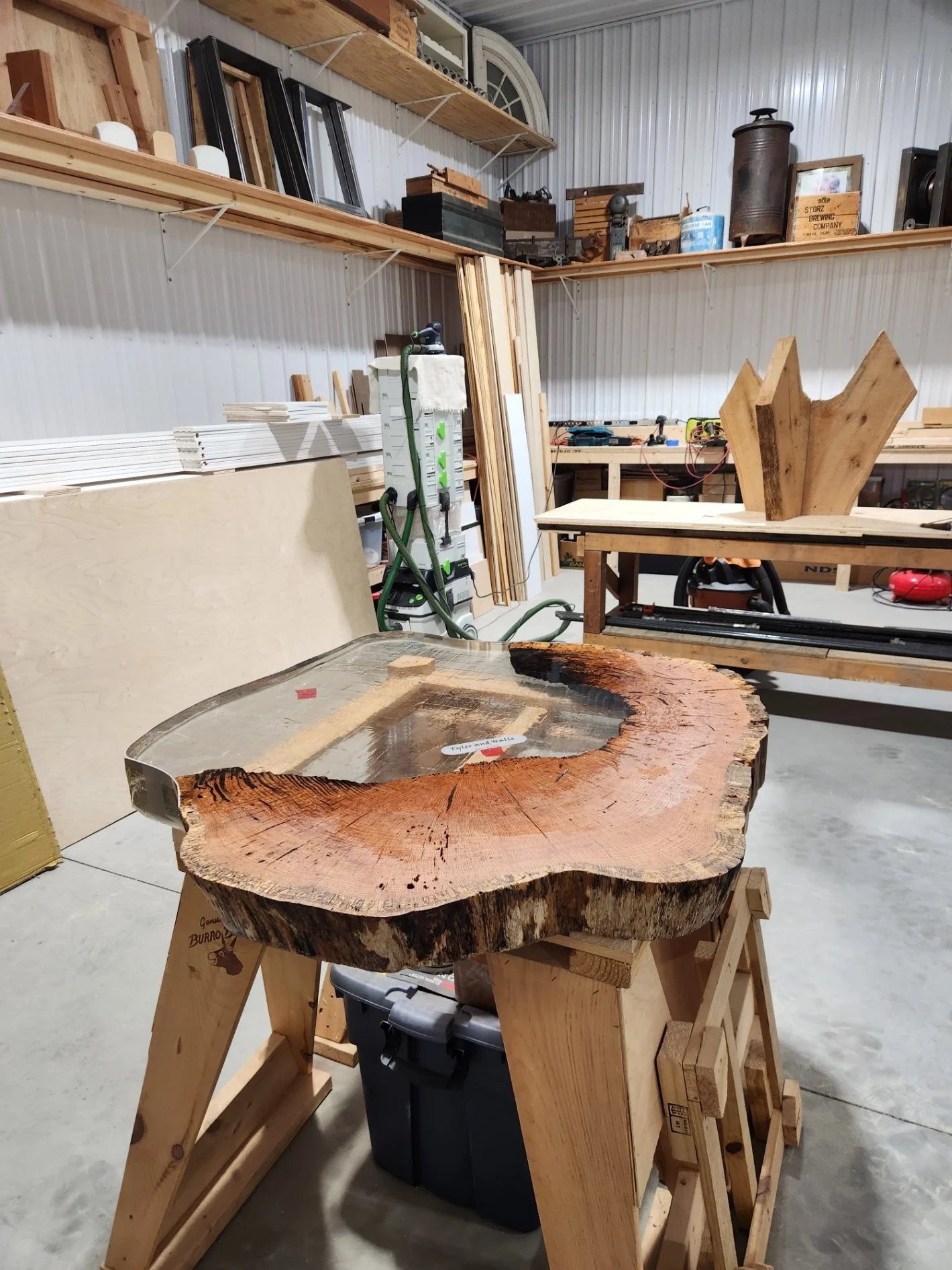 A woodworking workshop with various tools and wood pieces, featuring a large wood slab on a stand in the foreground.