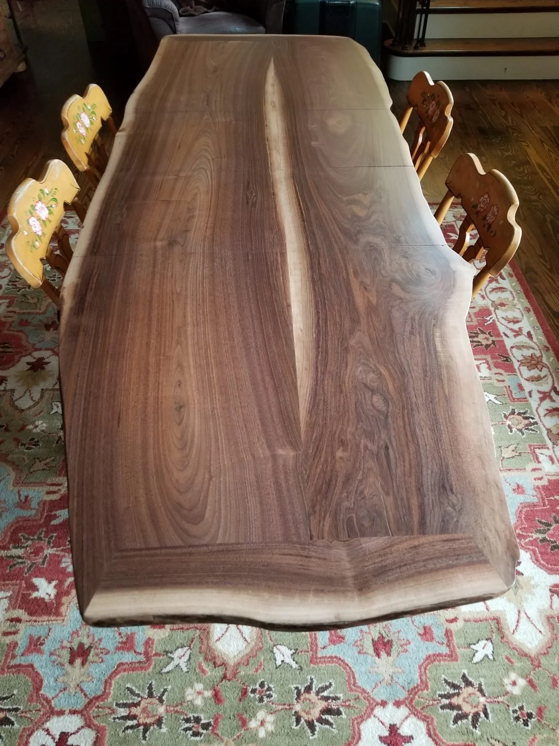 A large wooden dining table with a natural, live edge design, placed on a patterned area rug in a home setting, with four chairs around it.