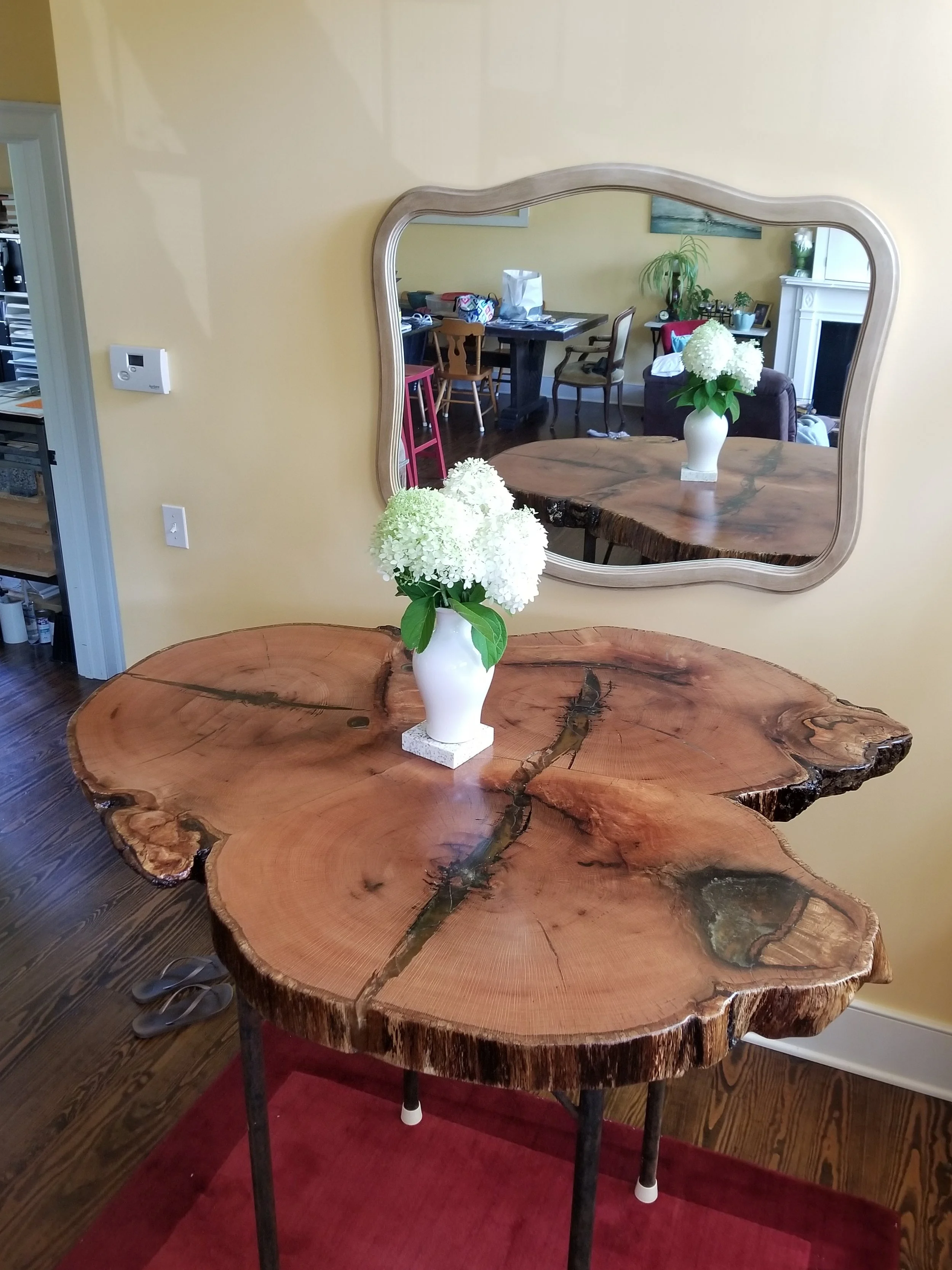 A rustic oak high top table with natural live edge, painted white flower vase with white hydrangeas, reflected in a wall-mounted mirror, in a cozy living area with yellow walls, a fireplace, and a dining room visible in the background.
