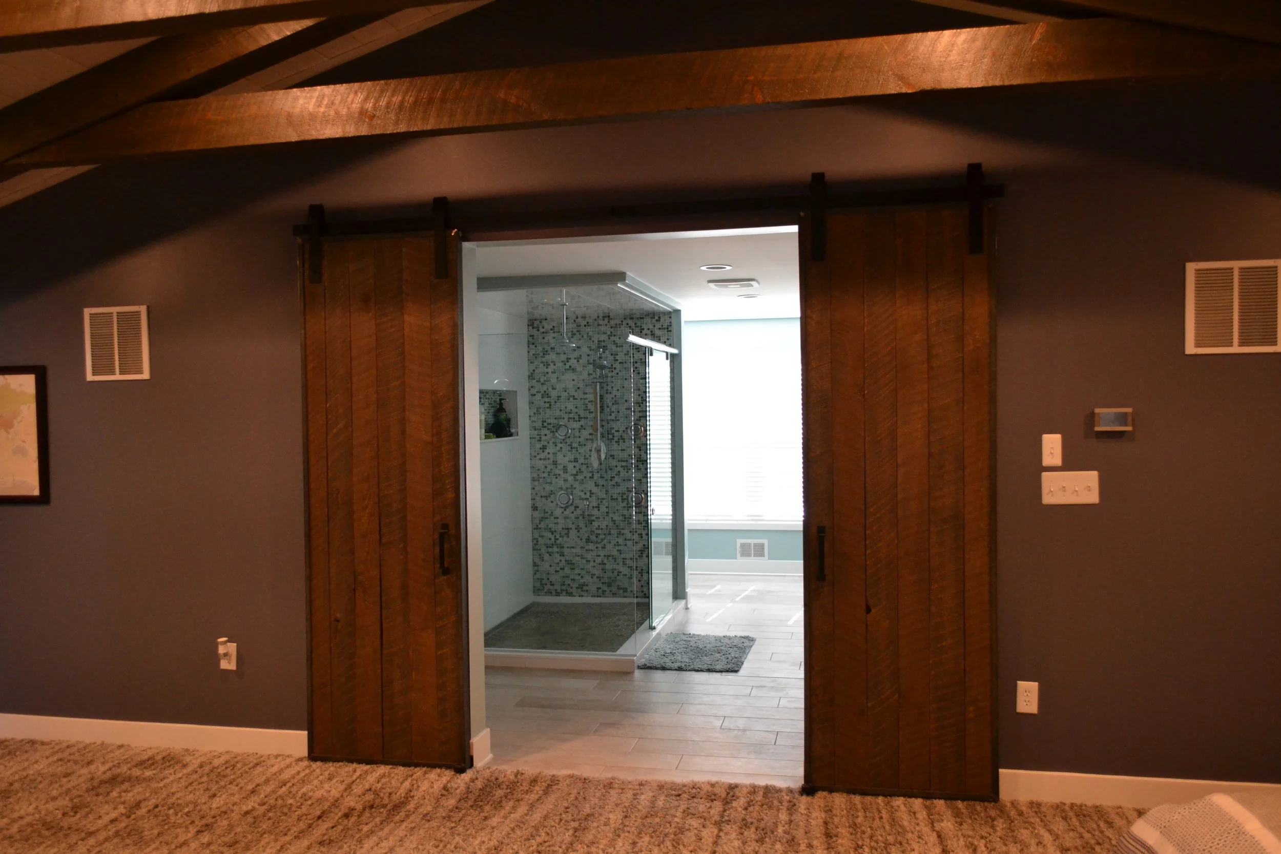 Interior view of a bedroom with carpeted floor, gray walls, and a wooden sliding barn door leading to a bathroom with a tiled shower.