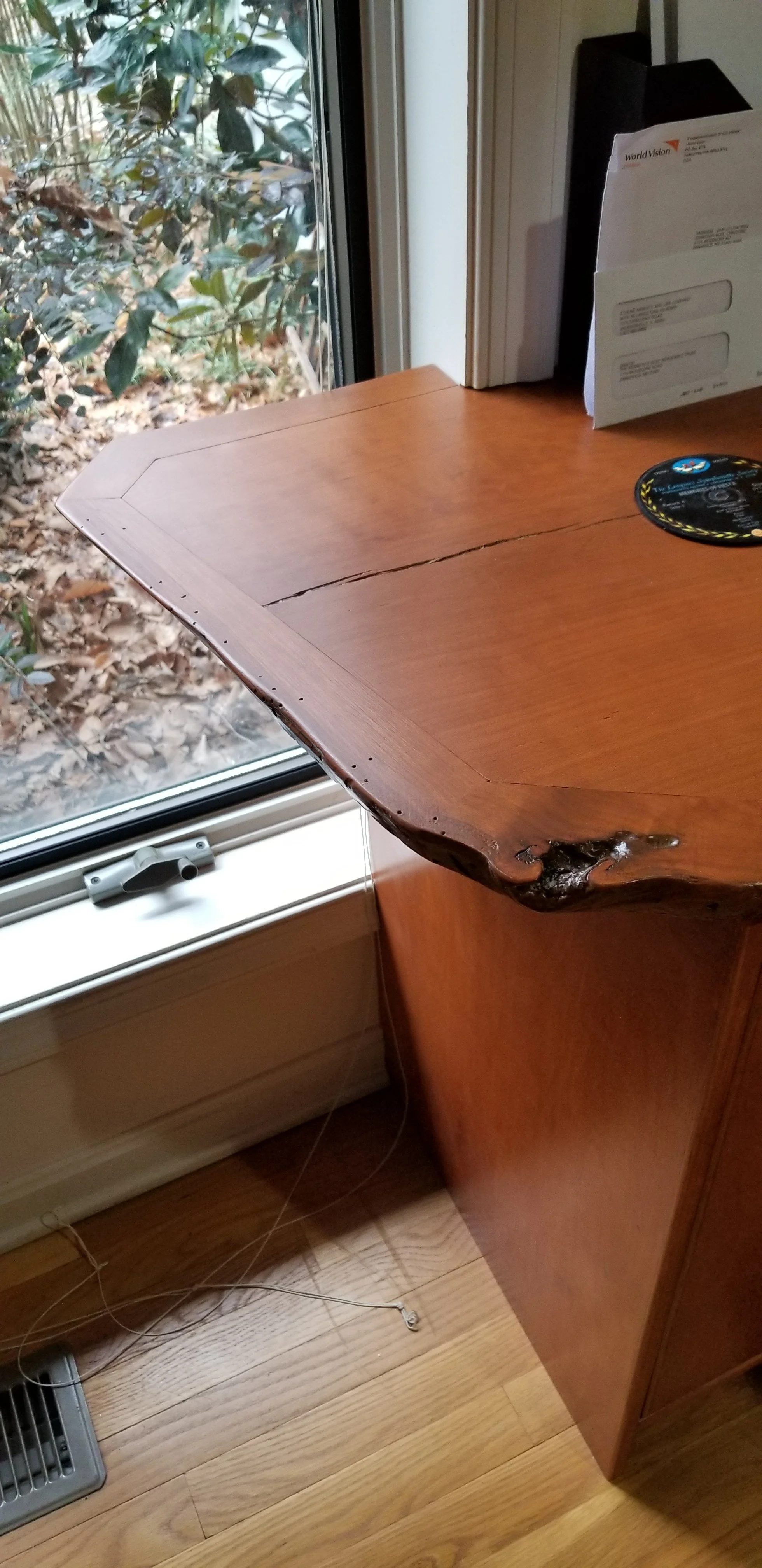 A damaged wooden table with a large crack and a broken edge near a window with a view of bushes.