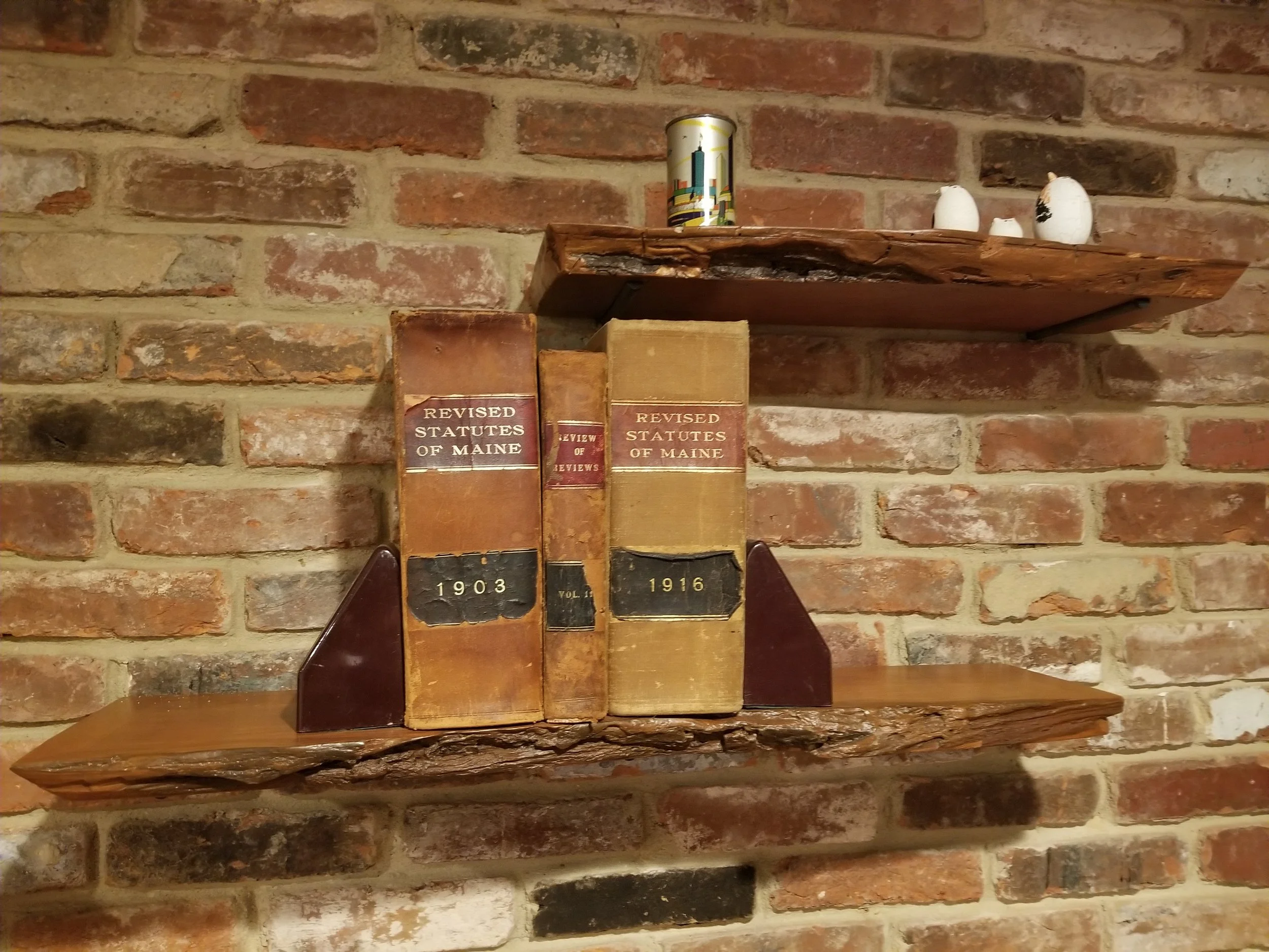A brick wall with cherry live edge shelves holding three bulbs, a cylindrical container with a cityscape design, and three old books titled 'Revised Statutes of Maine' aged from 1903 and 1916.
