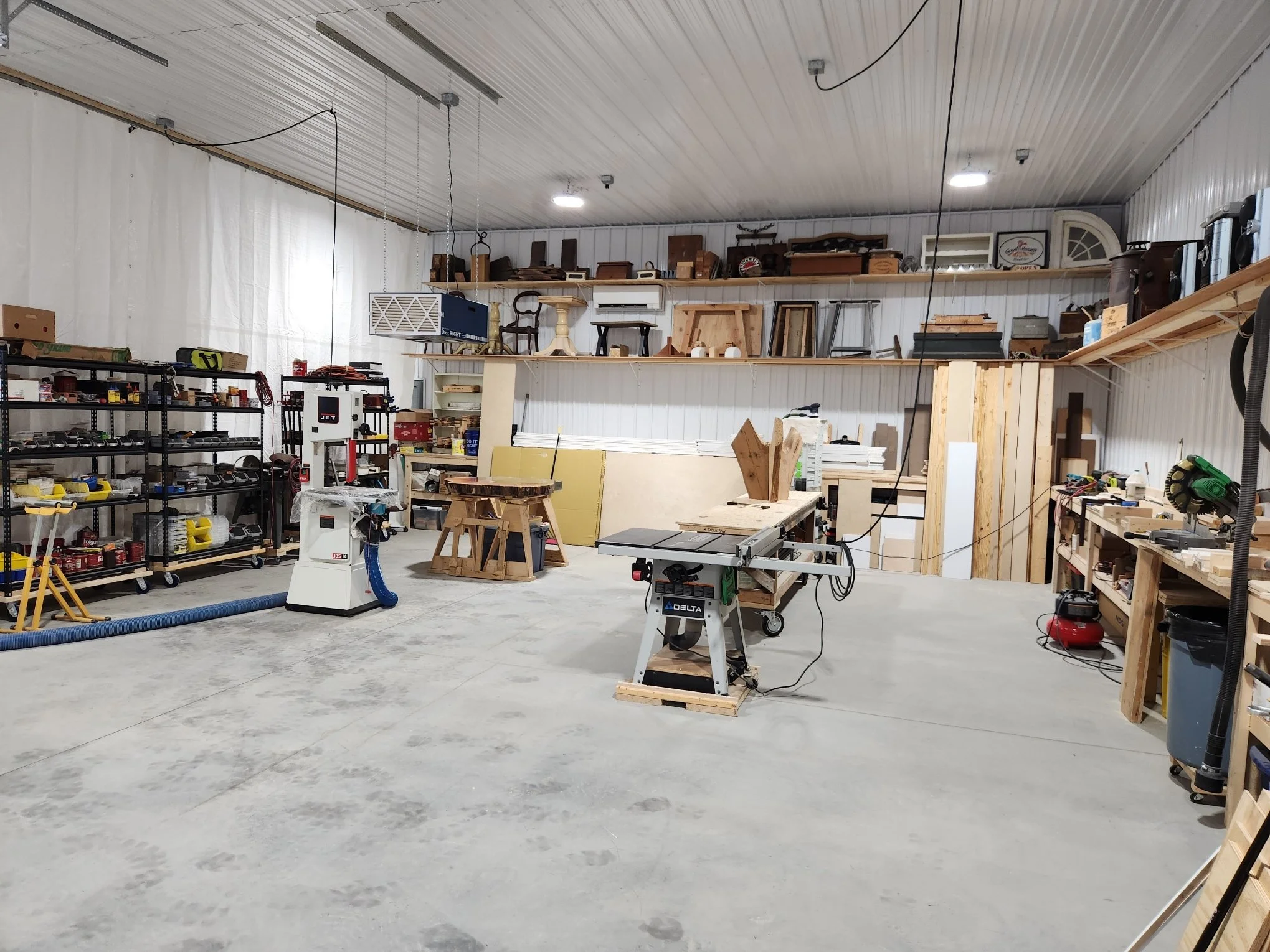 A spacious workshop with shelves filled with tools, equipment, and wooden materials. Various workbenches and woodworking machines are also visible, with a concrete floor and a ceiling with led lights.
