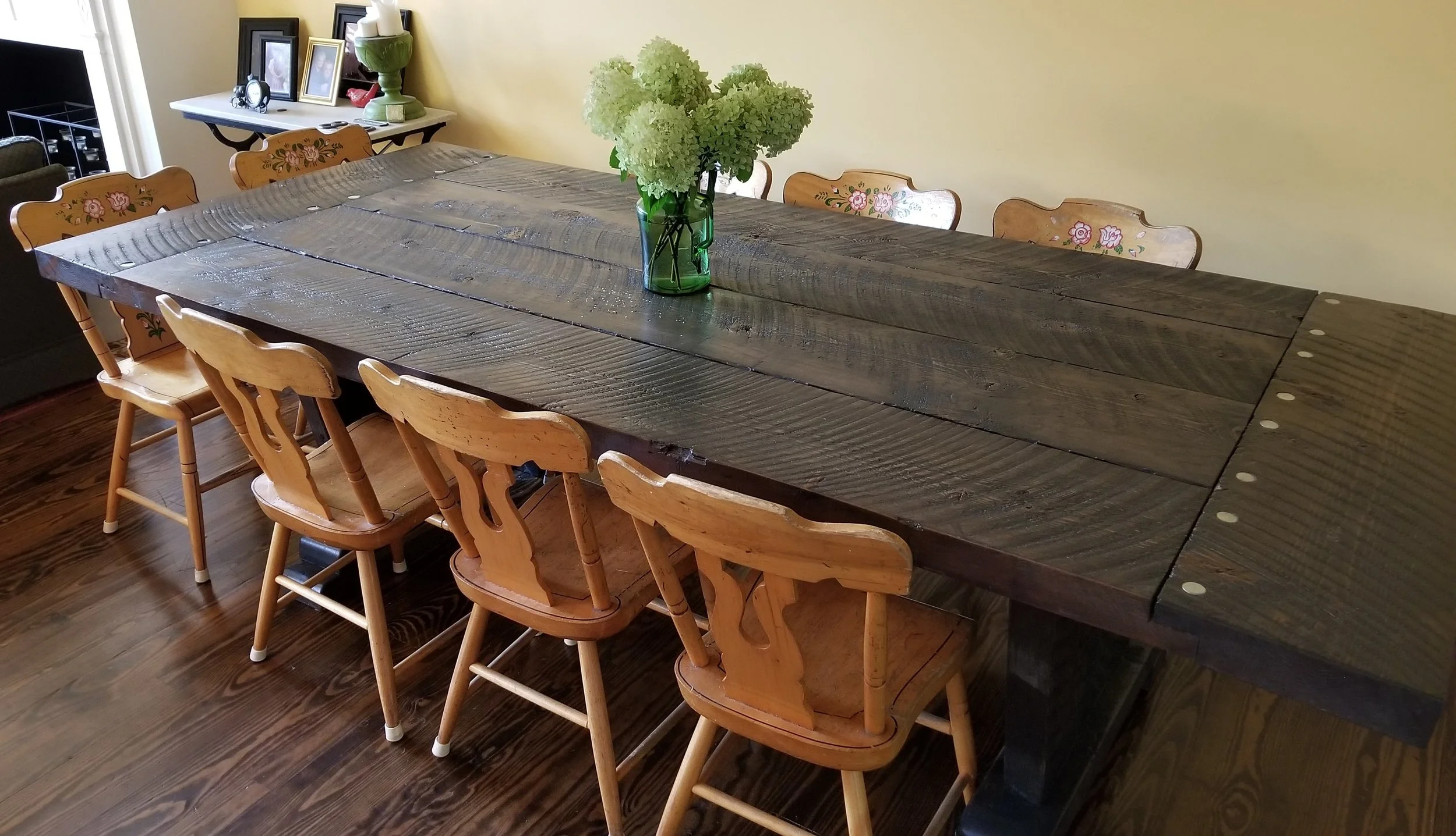 A wooden dining table with a dark finish in a room with hardwood floors. A green vase with white hydrangeas is in the center of the table. Six wooden chairs with floral painted designs on the backrests surround the table. A sideboard with picture fra