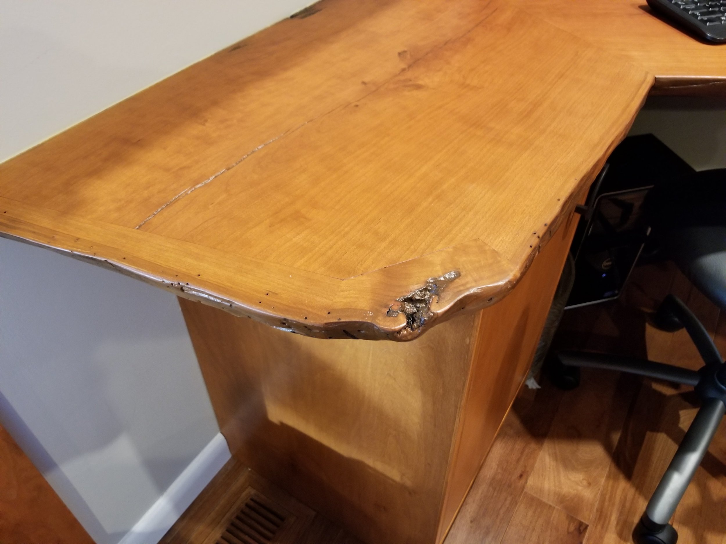 A wooden desk with a noticeable crack on its surface, showing some charred or burnt areas near the edge, with a portion of a keyboard visible in the top right corner, and a heating vent or grille at the bottom left.