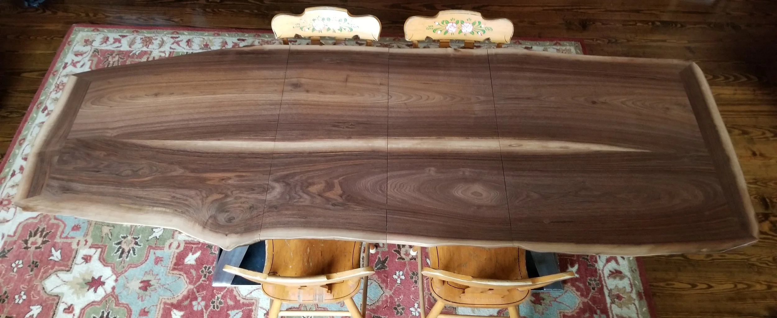A large walnut dining table with 2 book matched leaves, surrounded by four matching wooden chairs with curved backrests, placed on a patterned area rug on a wooden floor.