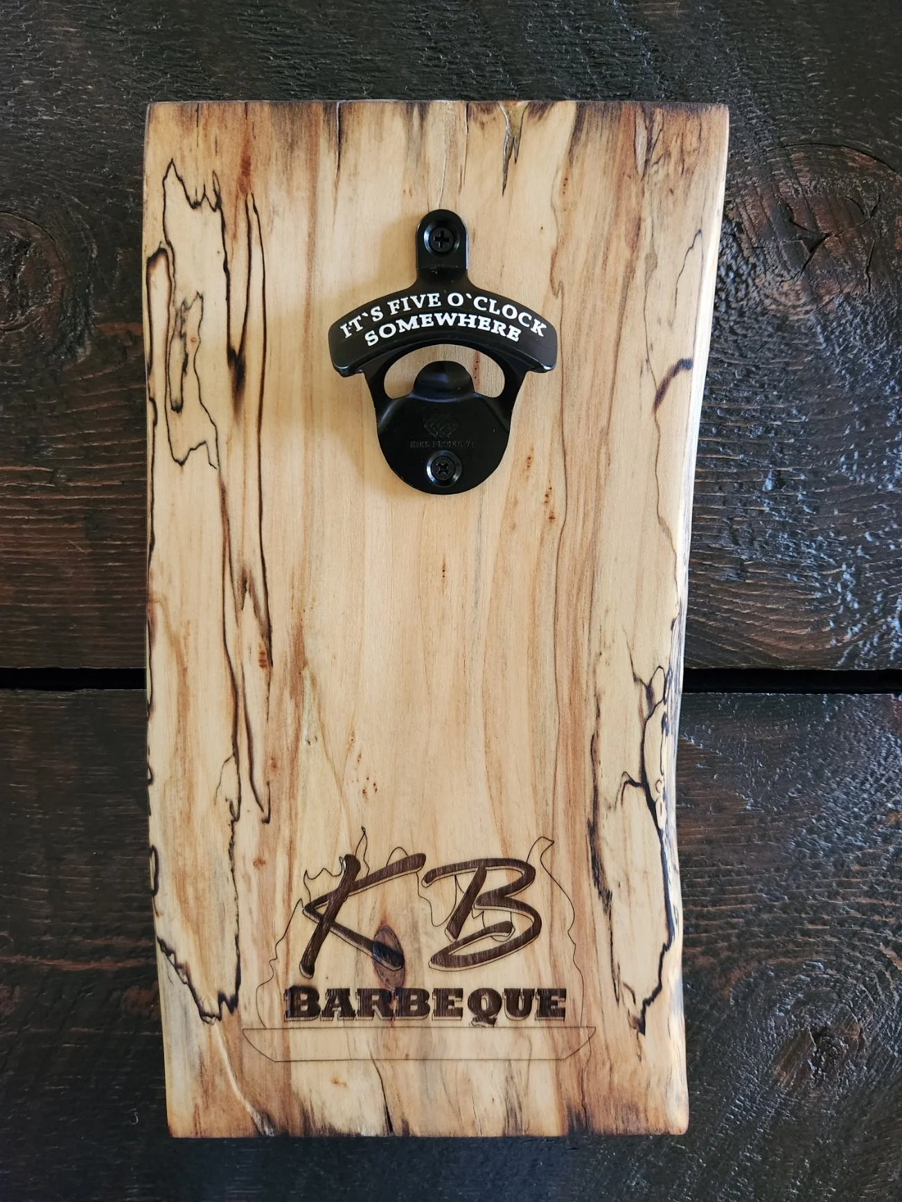 A wooden plaque with a mounted bottle opener and the phrase "It's five o'clock somewhere." The plaque has a natural wood grain pattern and could be be part of a bar or barbecue setup.