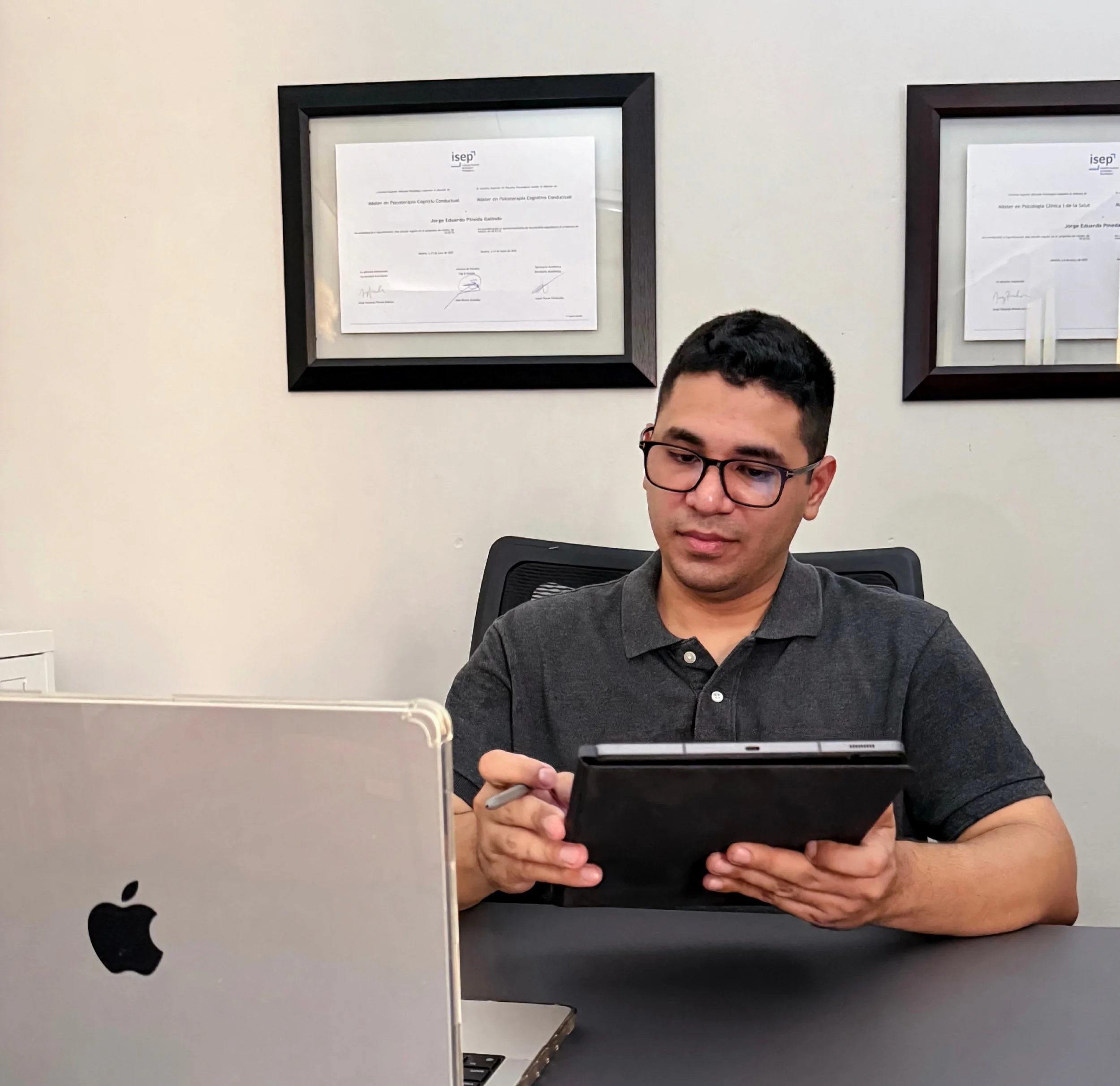 Jorge Pineda, terapeuta De San Pedro Sula, dando terapia psicológica en línea, está sentado frente a su escritorio, con una iMac, sosteniendo una tableta. En el fondo se observan dos maestrías enmarcadas en la pared.