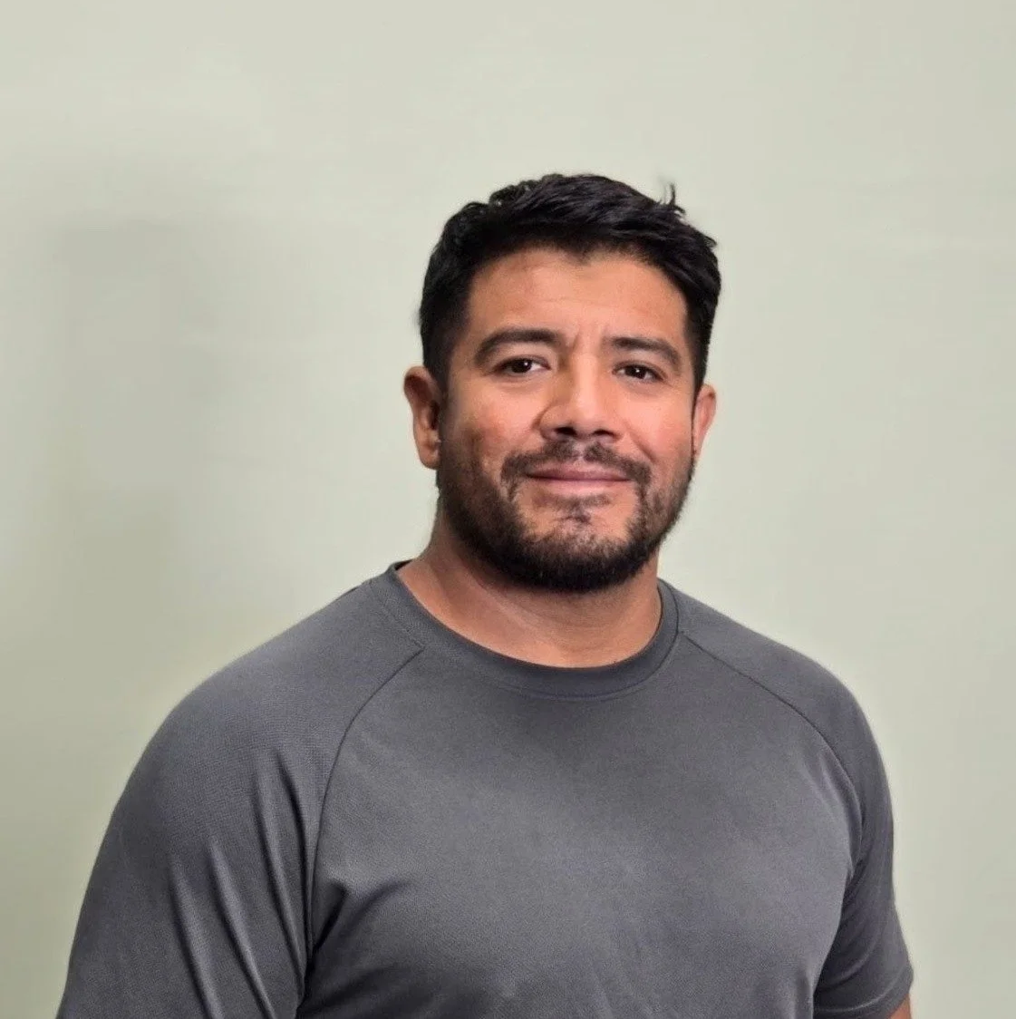 A man with dark hair and a beard wearing a gray t-shirt standing against a light-colored wall.