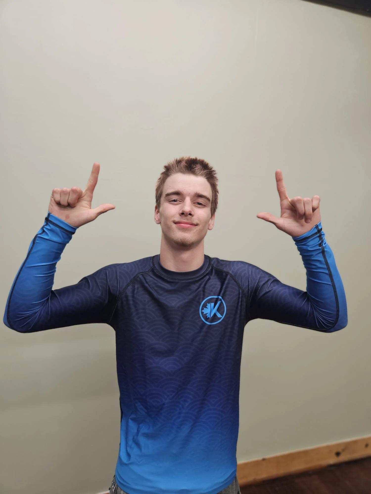 Young man smiling, wearing a black athletic shirt with blue long sleeves, standing against a plain light-colored wall, making a hand gesture.