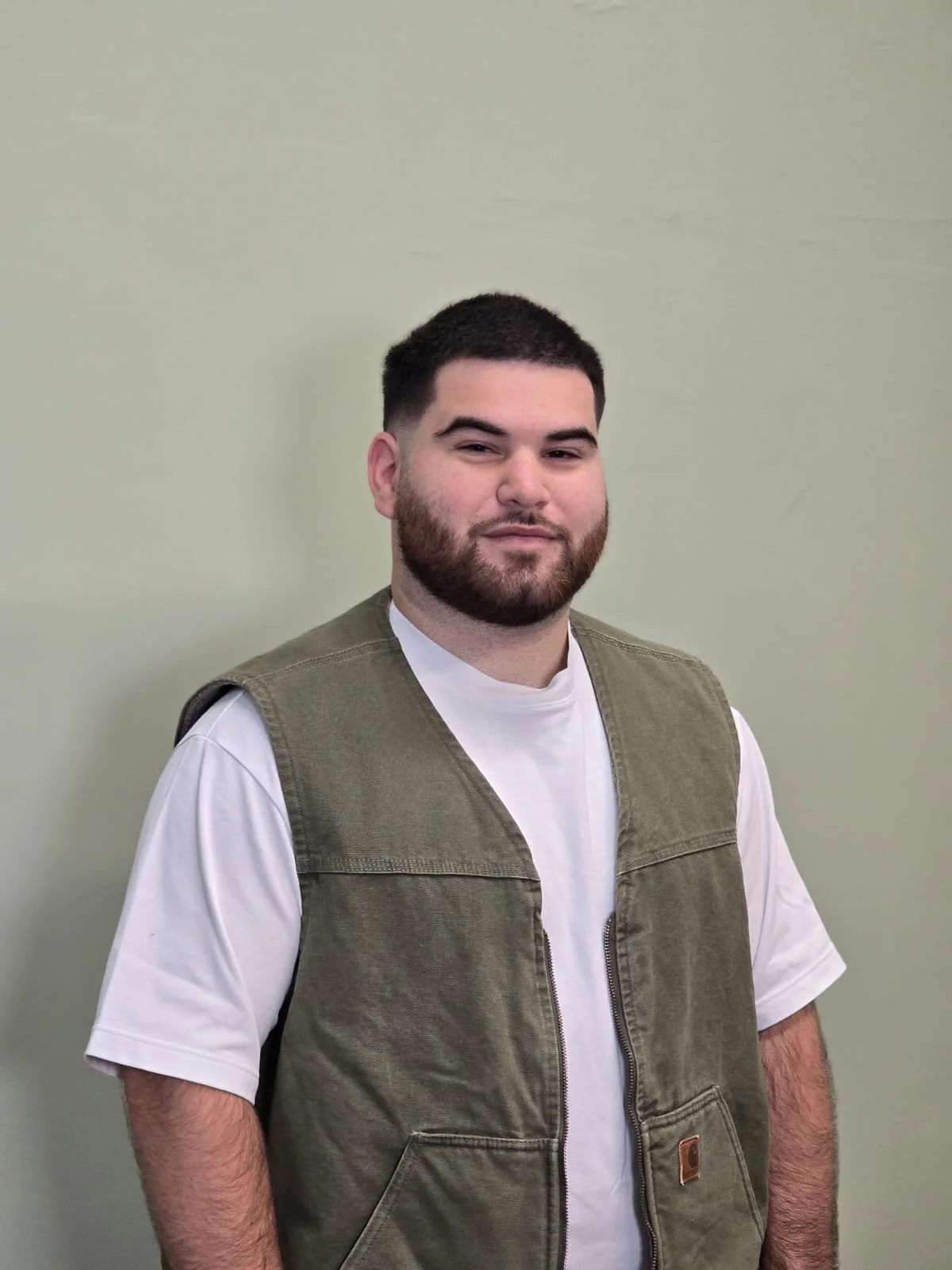 A man with dark hair and a beard standing against a plain light green wall, wearing a white t-shirt and an olive green vest.