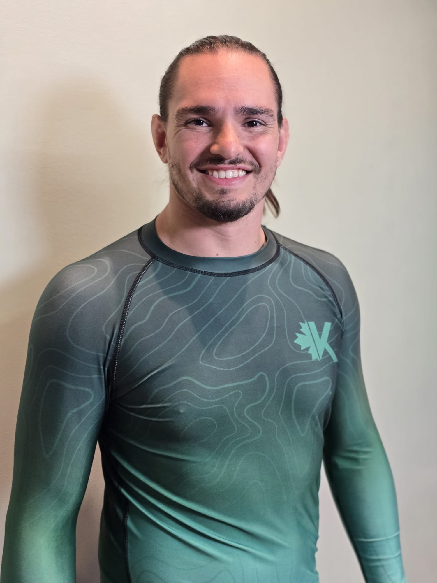 A smiling man with brown hair, beard, and mustache wearing a dark athletic shirt with a leaf logo.