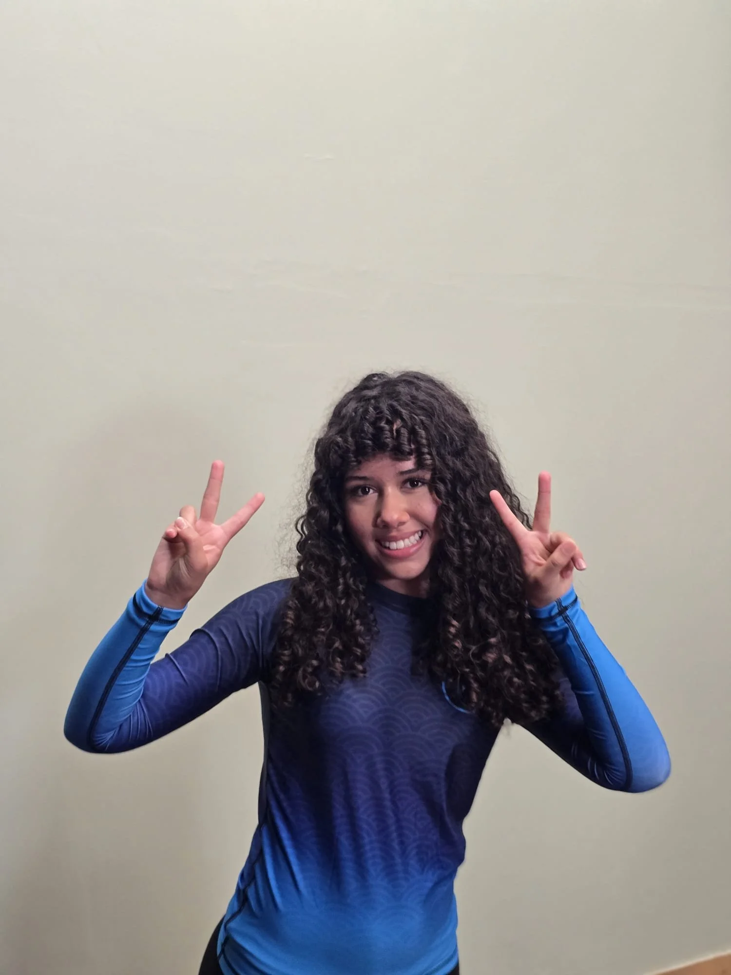 A young woman with curly dark hair smiling and making peace signs with both hands in front of a plain light-colored wall.
