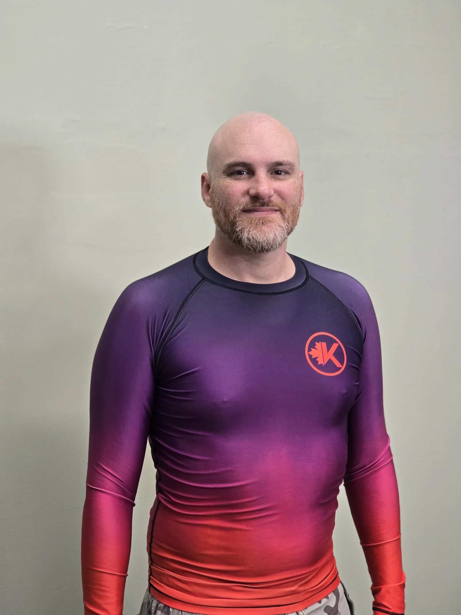 A man with a beard and shaved head standing against a plain light-colored wall, wearing a long-sleeved athletic shirt that fades from purple to red and has a logo on the chest.