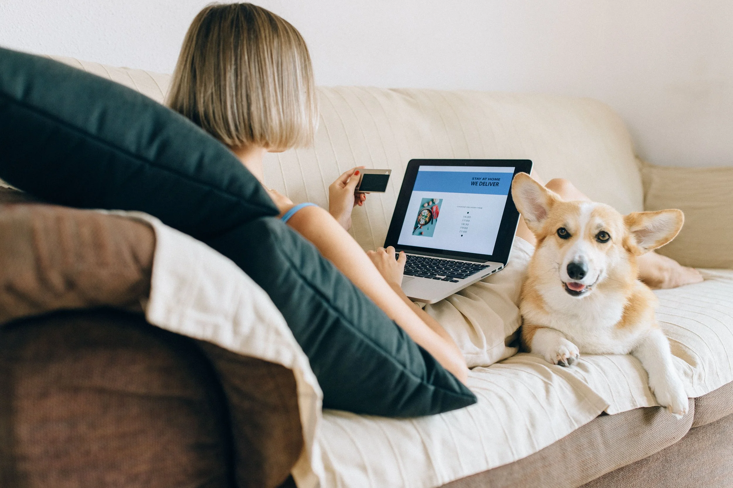 A pet business woman with blonde hair sitting on a beige couch with a dog laying beside her. She is holding a smartphone and using a laptop, which displays a webpage about staying at home and delivery options.