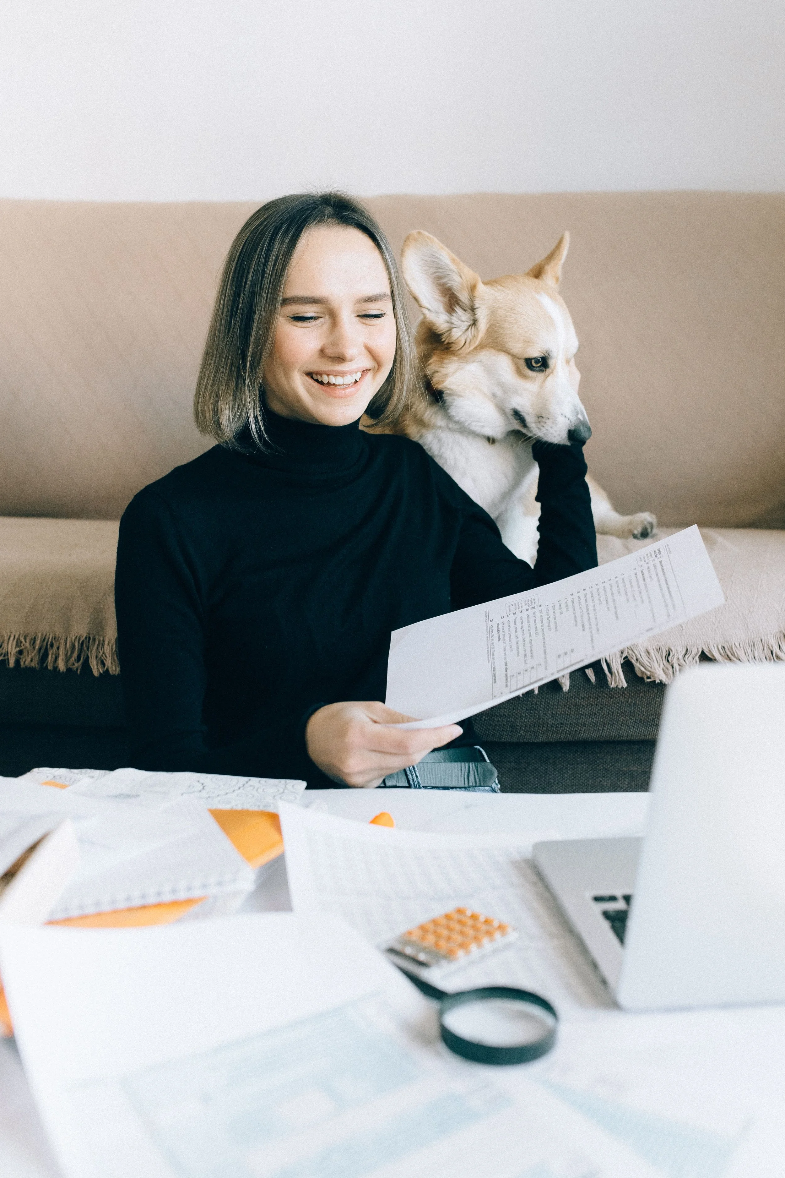A woman with shoulder-length hair and a black turtleneck shirt sitting on a beige couch, smiling and reading a paper. A dog with white and beige fur is sitting beside her, looking to the side. There are papers, a calculator, and a roll of tape on the table in front of her.