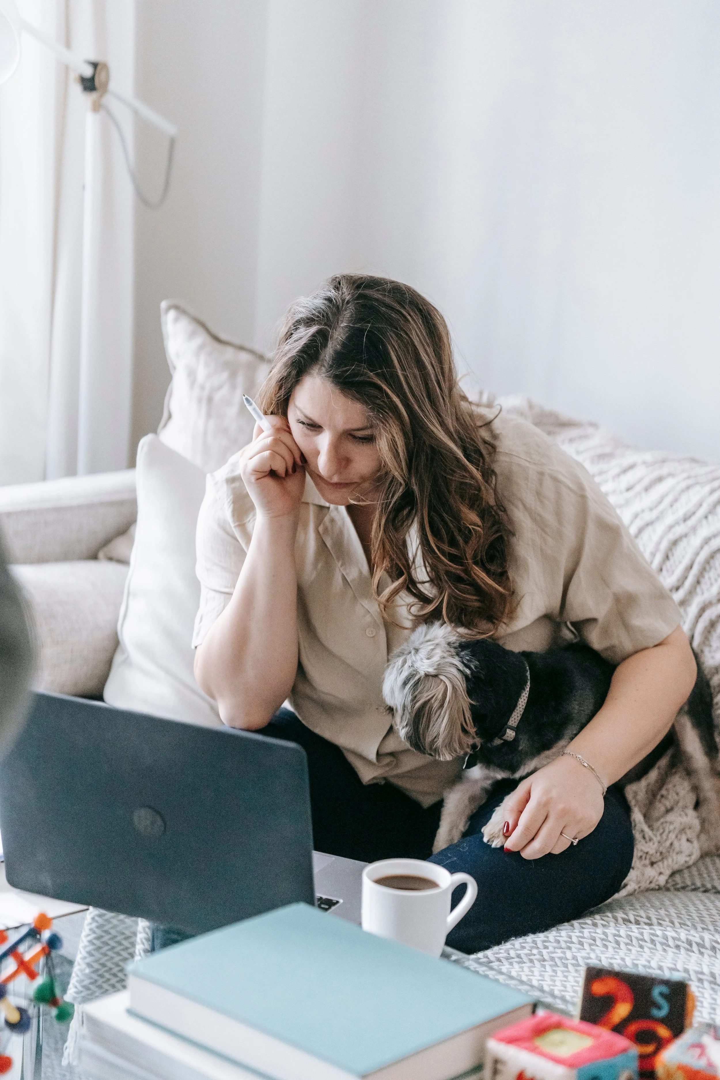 A woman with long wavy brown hair sitting on a couch, looking at a laptop, holding a small dog, with books, a cup of coffee, and colorful toys on the table in front of her in a cozy, well-lit living room.