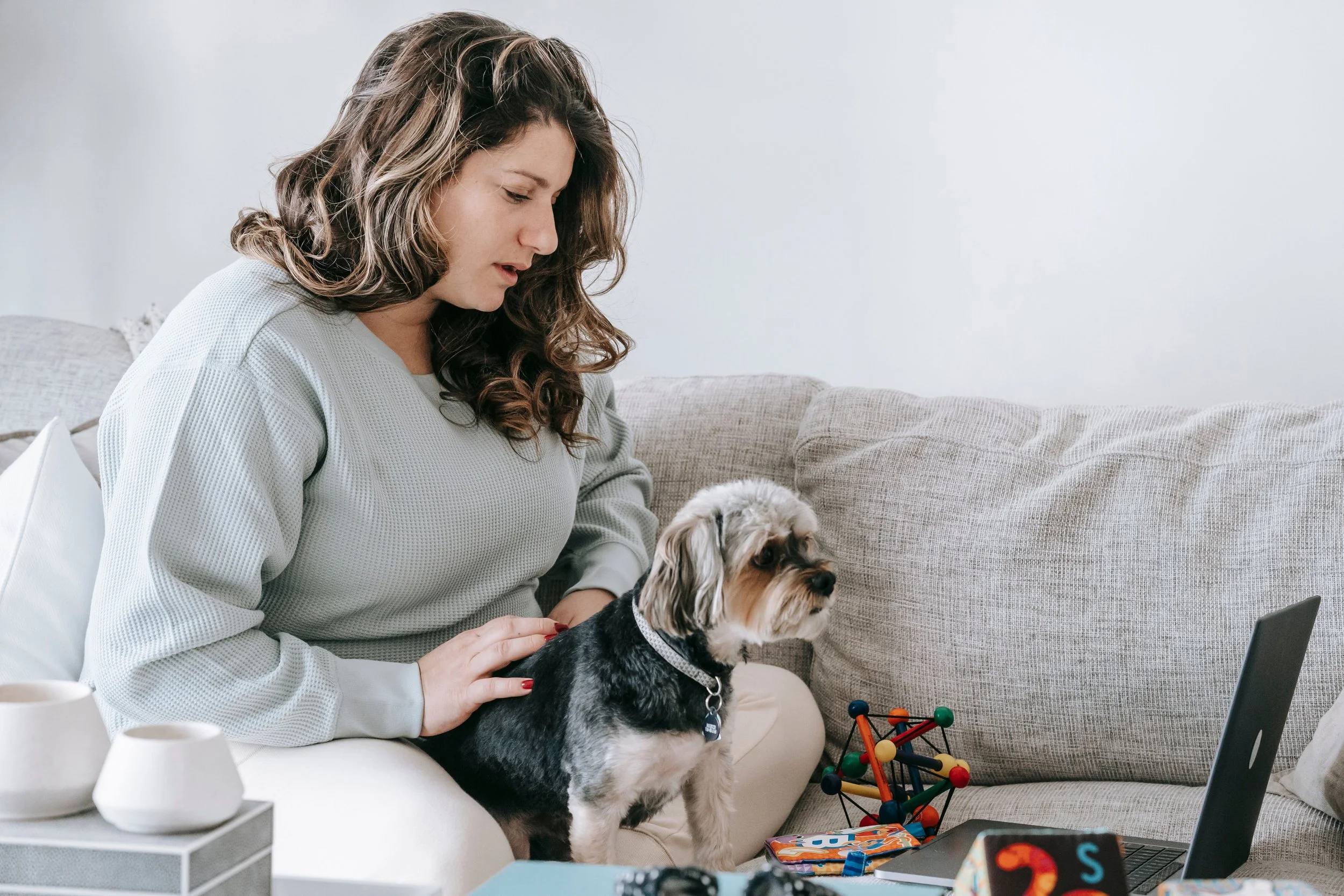 A woman with brown wavy hair, wearing a light-colored sweater, sitting on a couch with a small dog and working on a laptop. The background includes a beige sofa and a white wall.