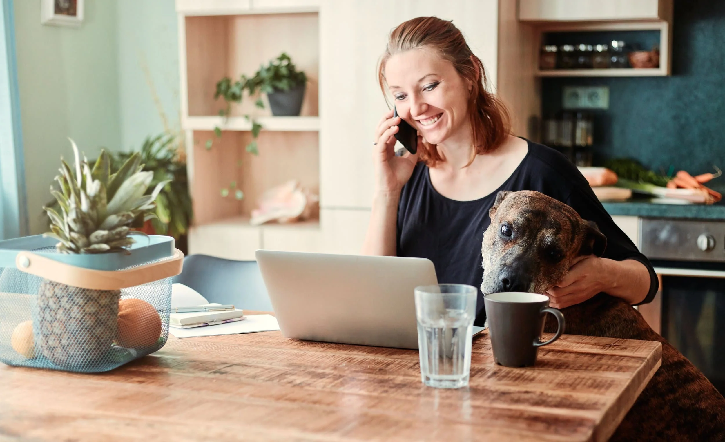 A dog walker with red hair smiling on the phone while holding a brown dog, sitting at a wooden table with a laptop, glass of water, coffee mug, and a pineapple in a basket, in a cozy kitchen.