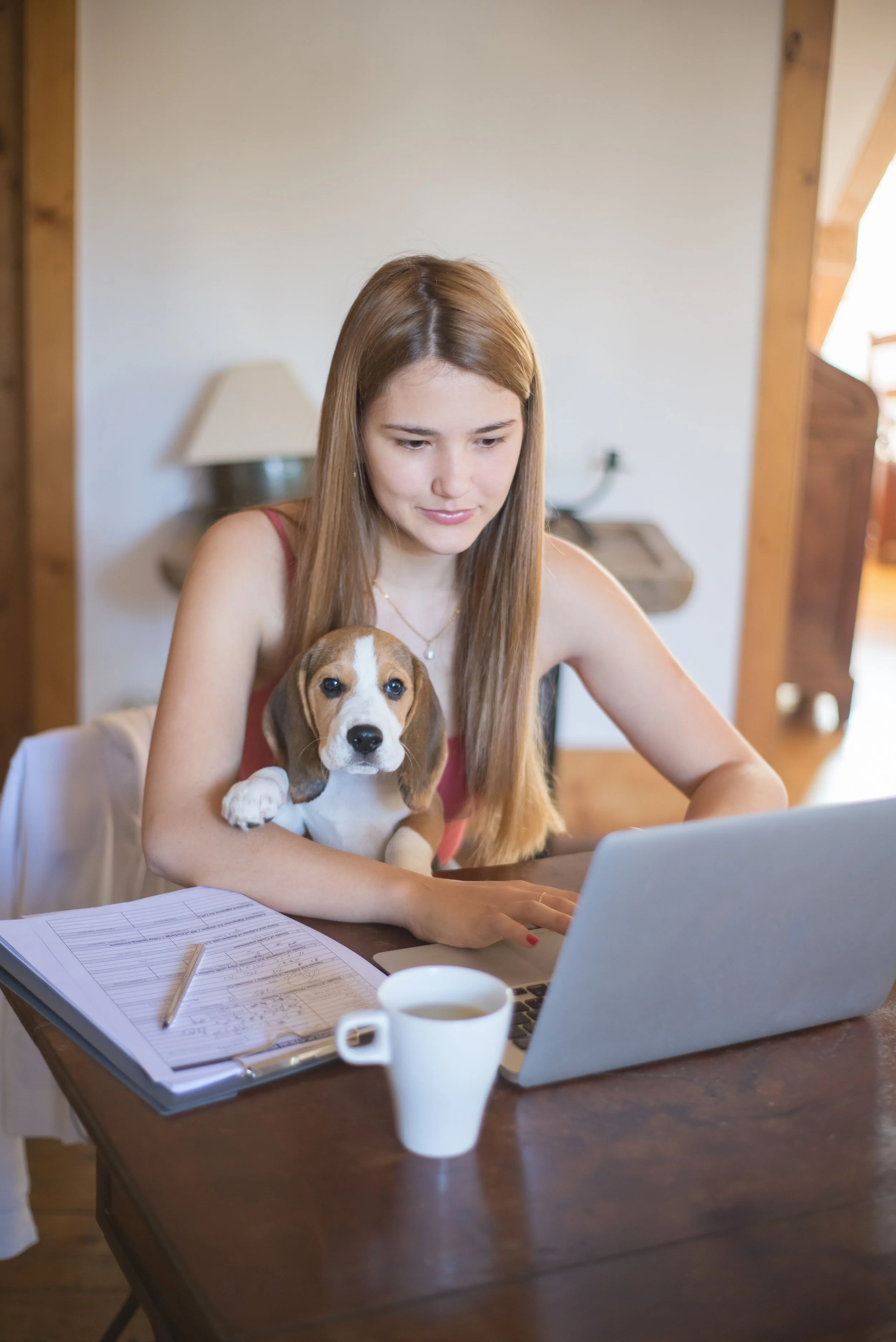 A dog walker with long reddish-brown hair working on a laptop at a wooden table, holding a puppy, with notebooks, a pen, a coffee cup, and a lamp in the background.