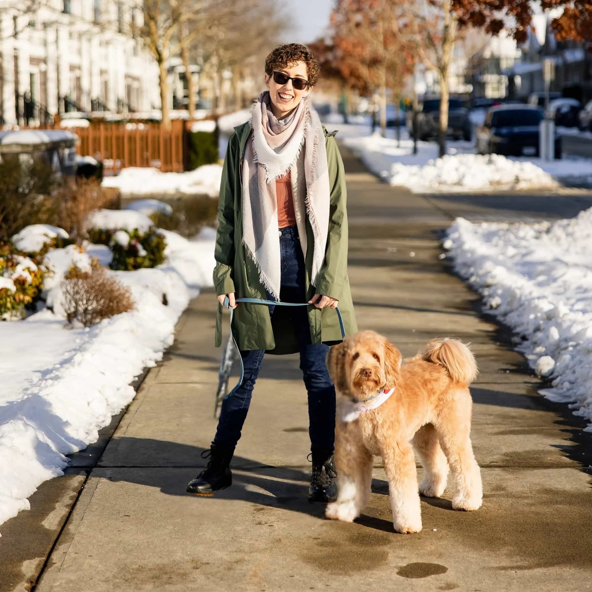 A woman walking two dogs on a snowy sidewalk in a residential neighborhood during daytime. She is smiling, wearing sunglasses, a green jacket, a scarf, and hiking boots.