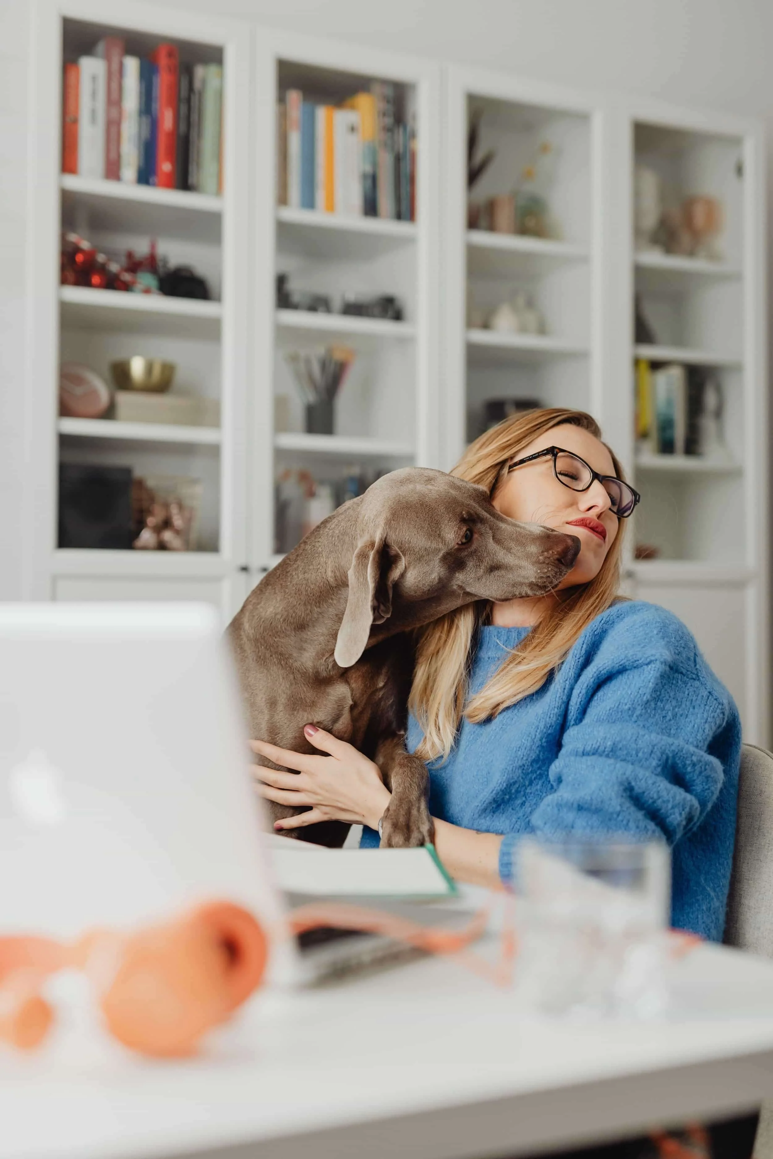 A woman in glasses sits at a desk, hugging a gray dog that is licking her face, with bookshelves in the background.