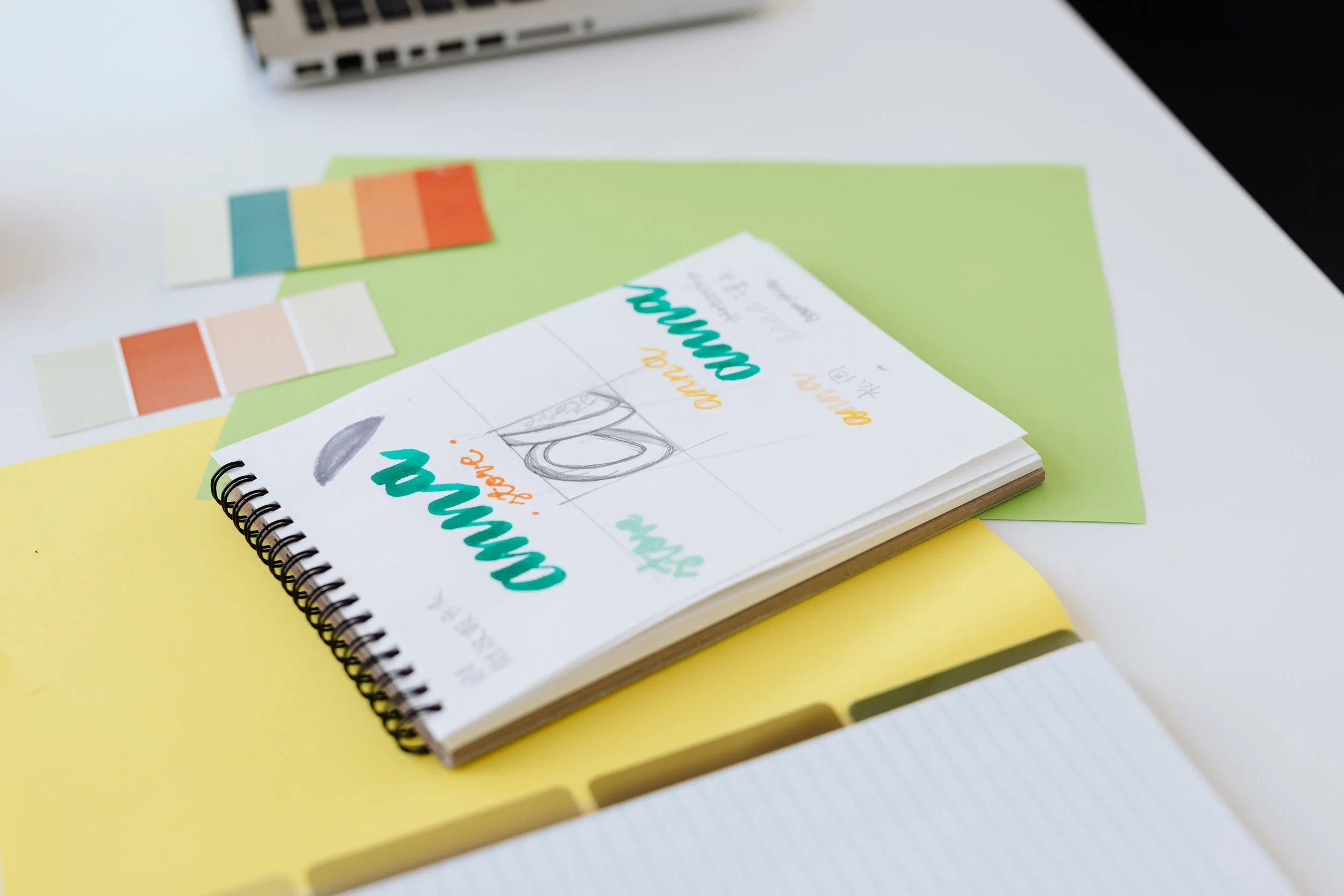 A spiral notebook with colorful handwritten text, next to color swatches, placed on a yellow and white surface.