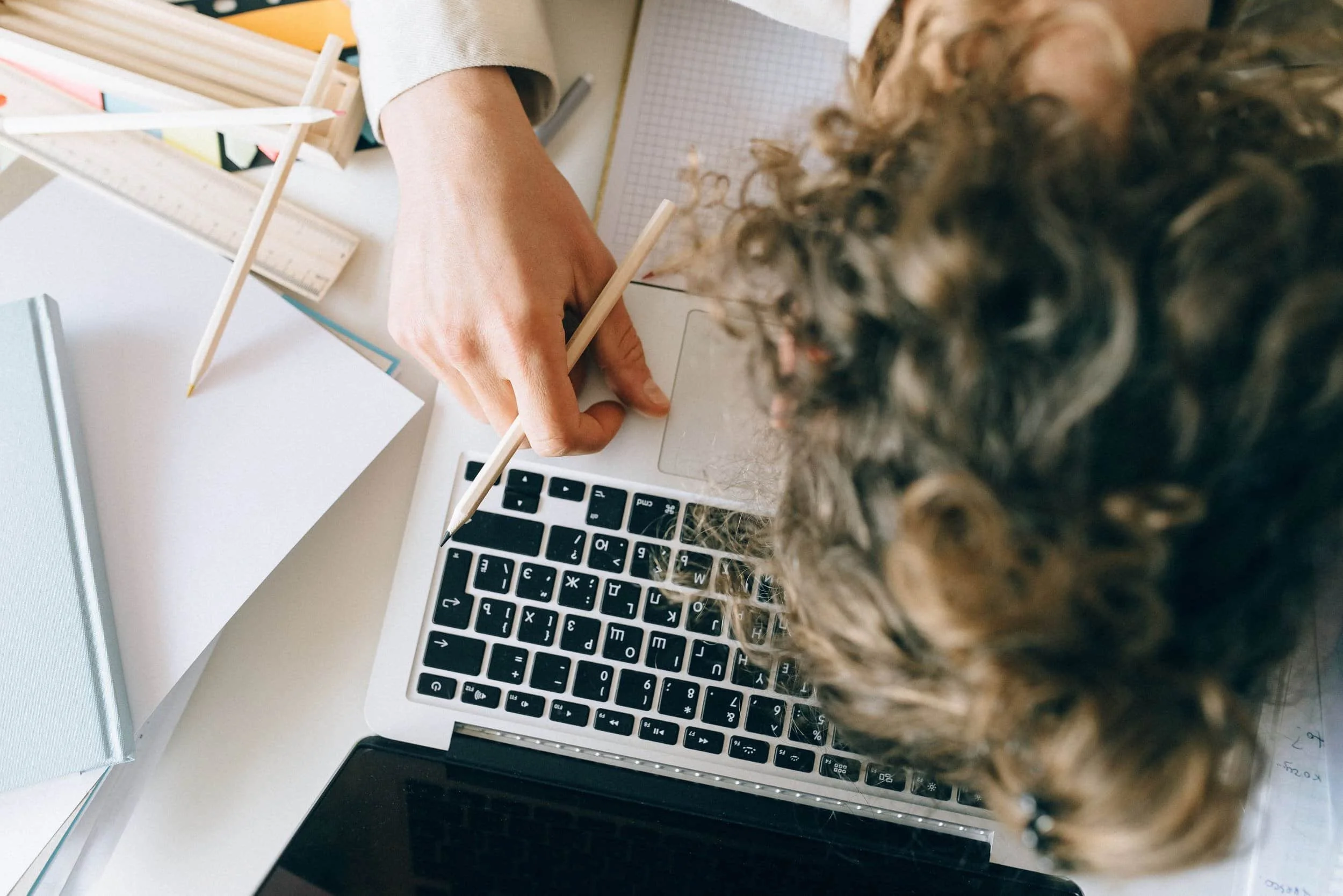 A person with curly hair working on a laptop surrounded by papers, a ruler, and a pen, with a notebook and a tablet nearby.