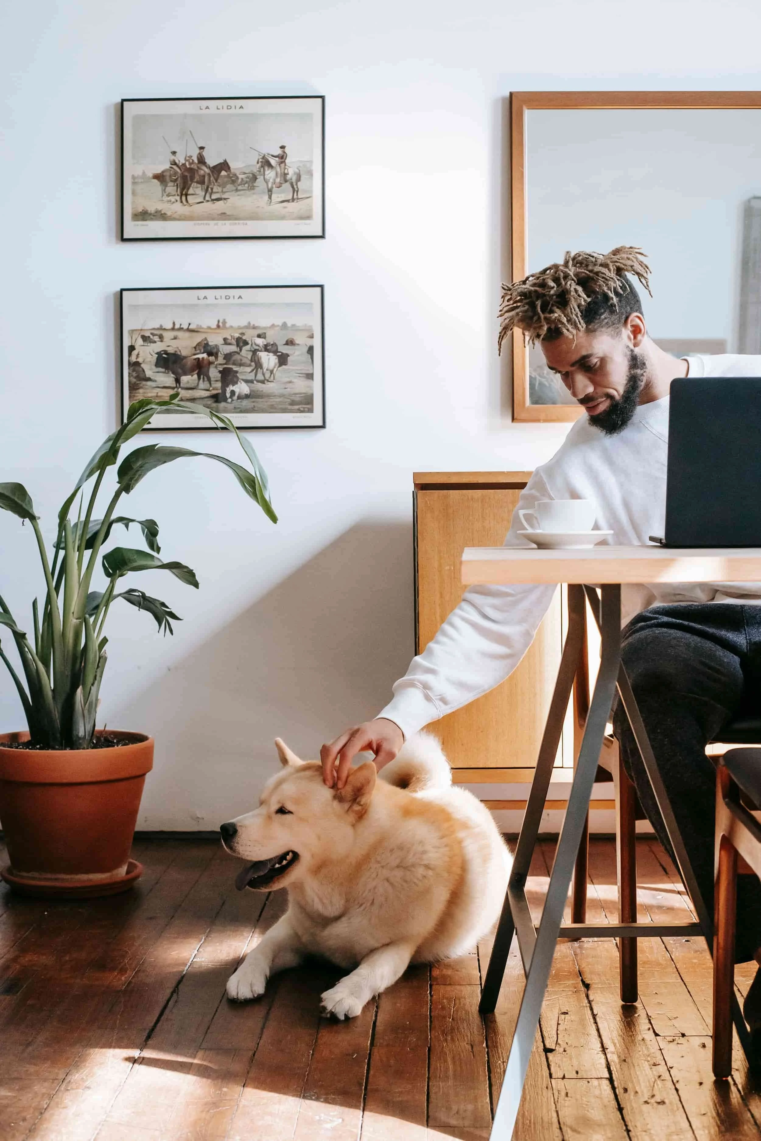 A man with distinctive dreadlocked hair and a beard is sitting at a wooden table, petting a large, fluffy, light-colored dog lying on the dark wooden floor. There is a potted plant on the floor and framed vintage photographs of cowboys and cattle on the white wall behind him. The man appears to be in a cozy room with sunlight streaming in.