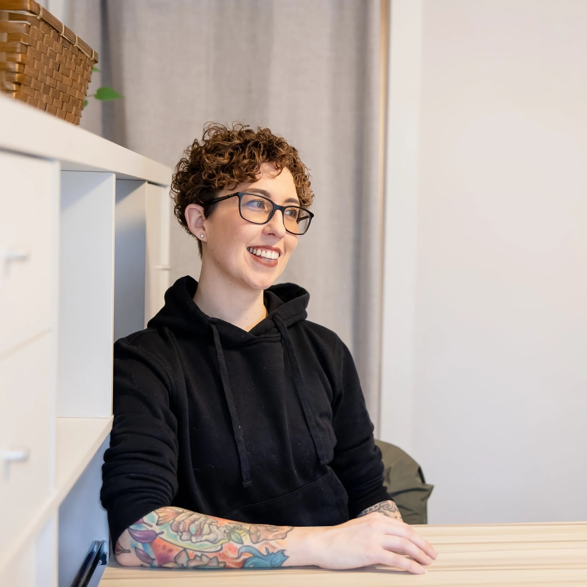 A young woman with short curly hair and glasses, smiling, sitting at a table with her arms crossed and decorated with colorful tattoos, in a modern interior space.