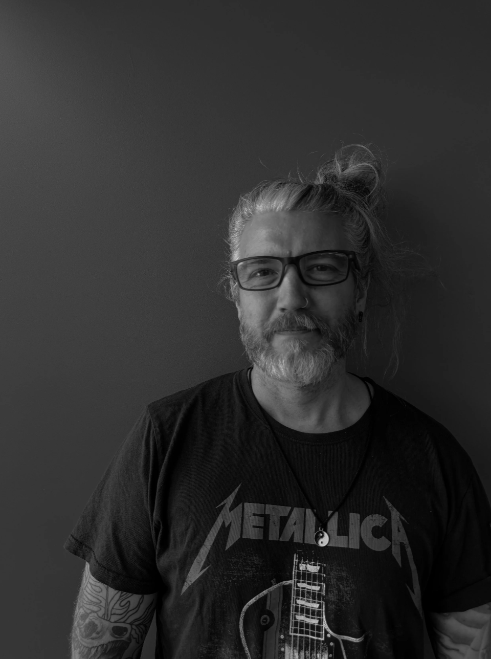 A man with long hair tied up in a bun, glasses, a beard, and a mustache, standing against a plain wall. He is wearing a Metallica T-shirt and a necklace with a yin-yang pendant.