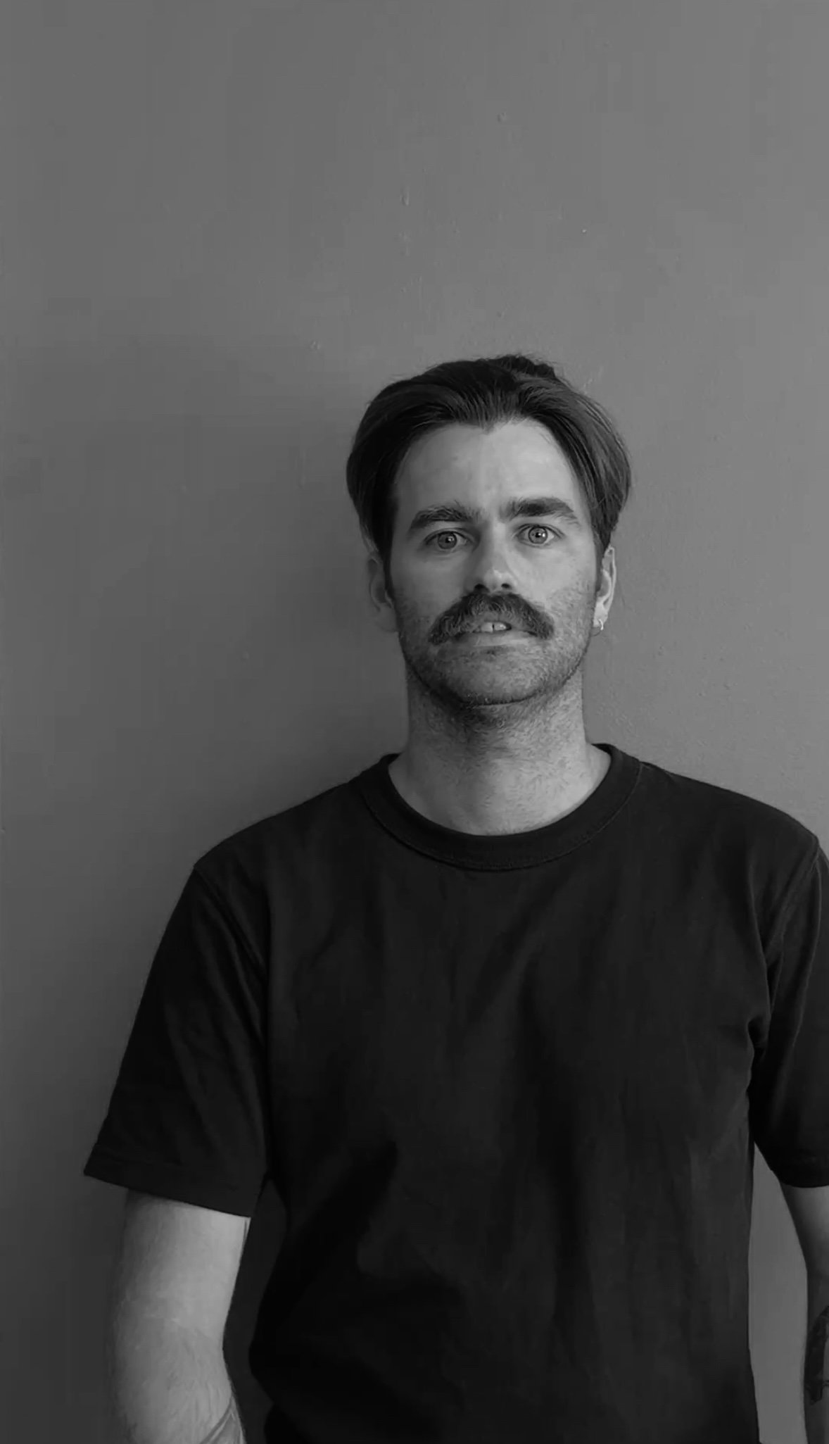 Black and white photo of a man with a mustache, wearing a dark t-shirt, standing against a plain wall.