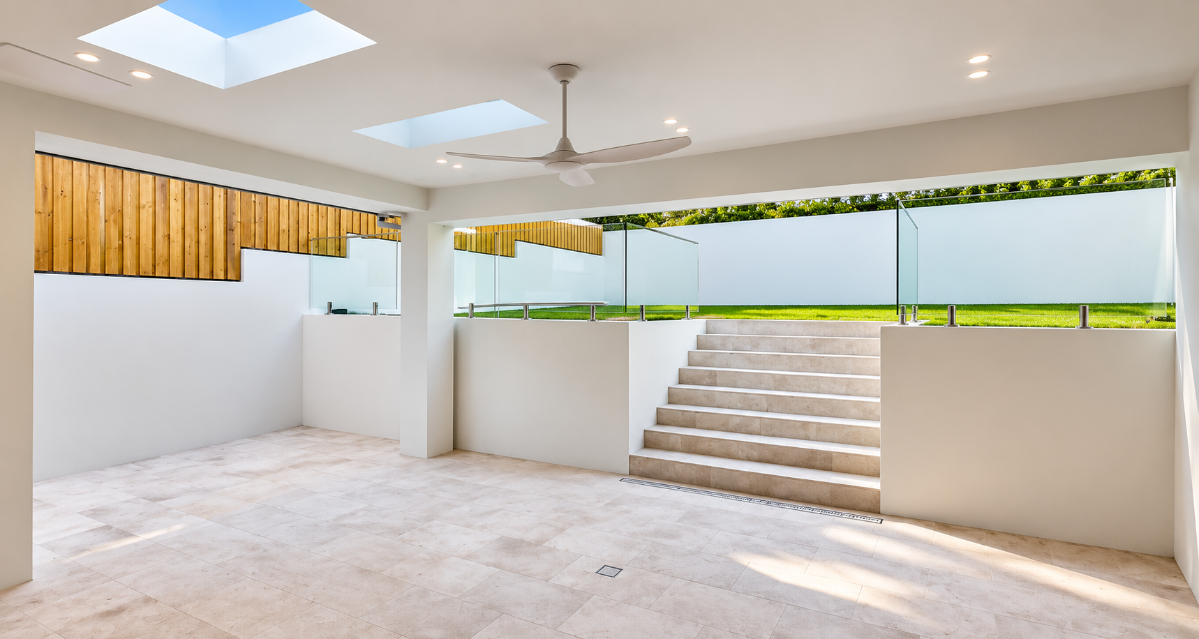 Interior view of a modern, minimalistic room with white walls, beige tile flooring, a ceiling fan, skylights, and a glass barrier leading to an outdoor grassy area with steps.