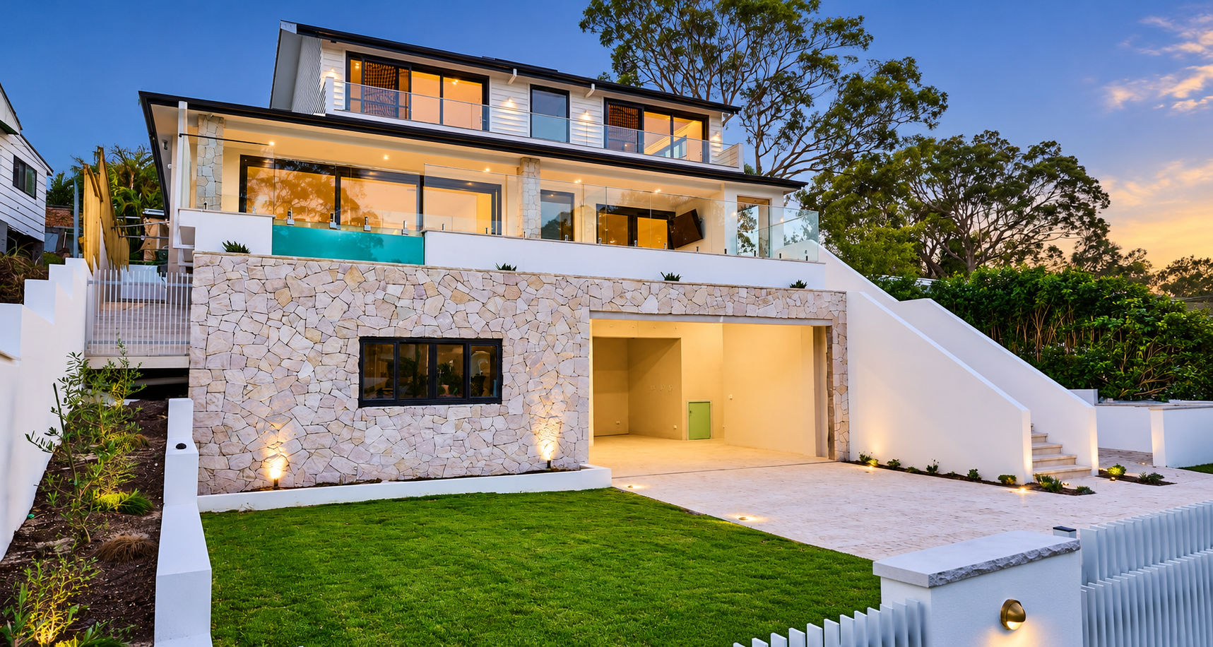 Modern multi-story house with stone facade, glass balcony railings, and outdoor lighting at sunset, with a well-maintained lawn and trees in the background.