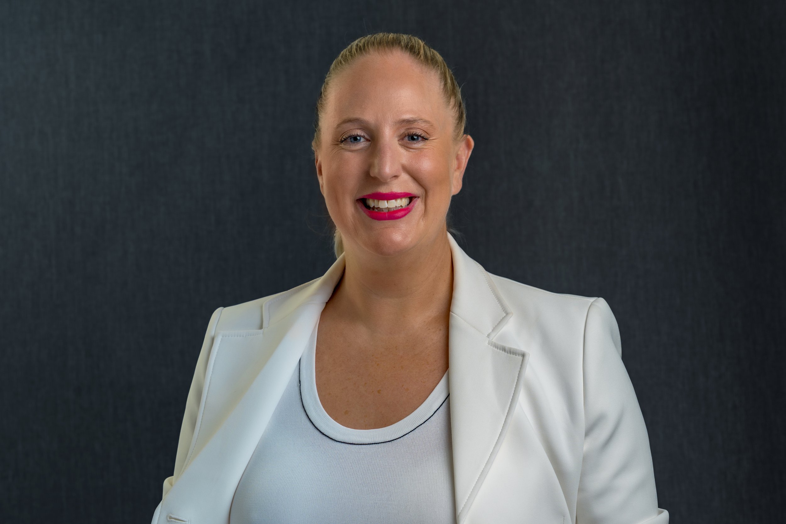 A woman with blonde hair in a ponytail, wearing a white blazer and a light-colored top, smiling against a dark background.