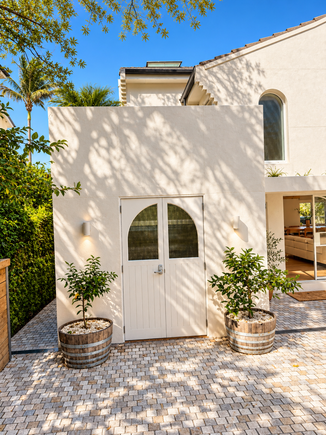 Modern white house with boxwood plants in wooden barrels and a cobblestone front patio under a clear blue sky.